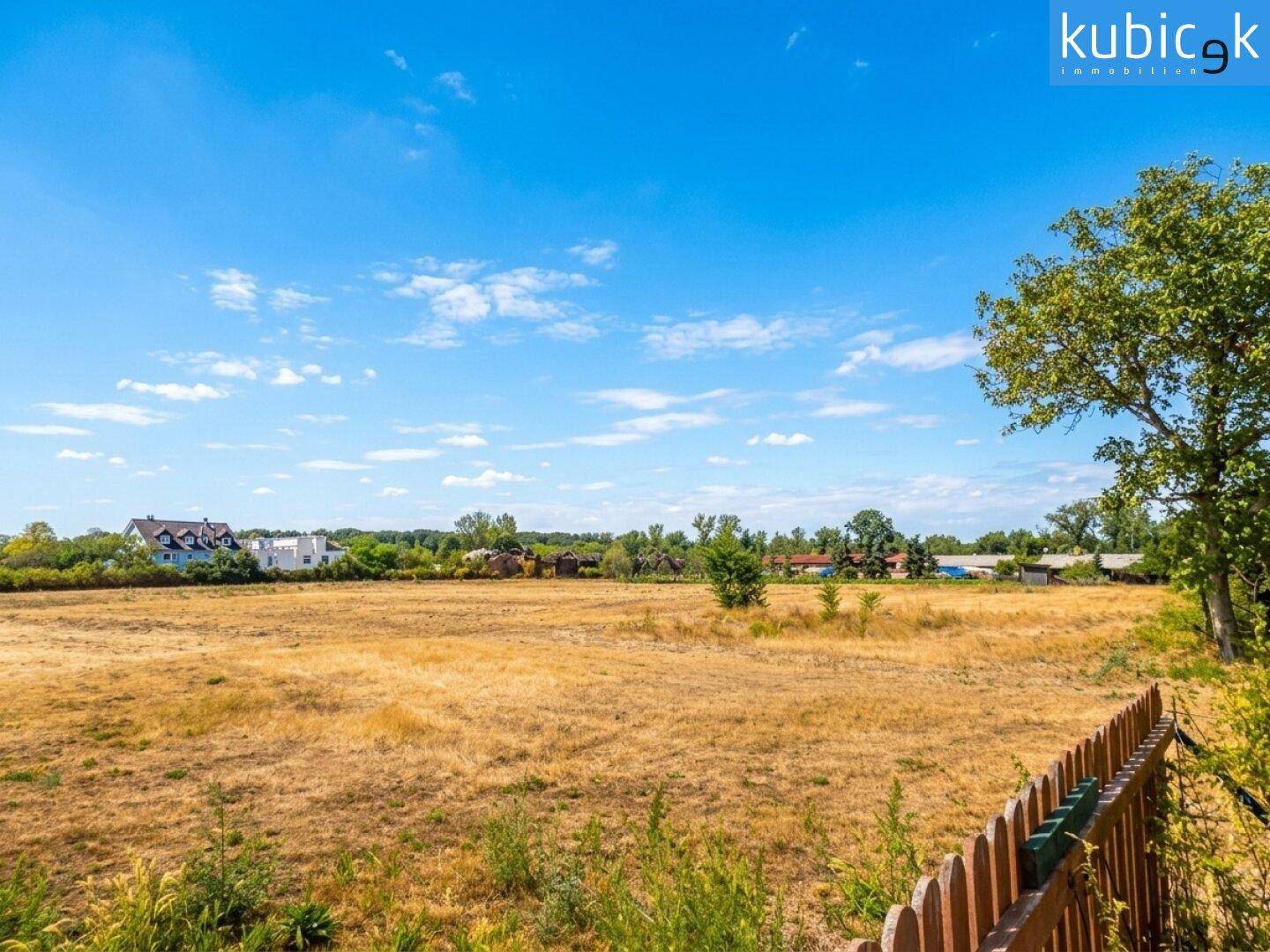 Weitläufige Landschaft mit trockenem Feld und Häusern in der Ferne unter blauem Himmel.