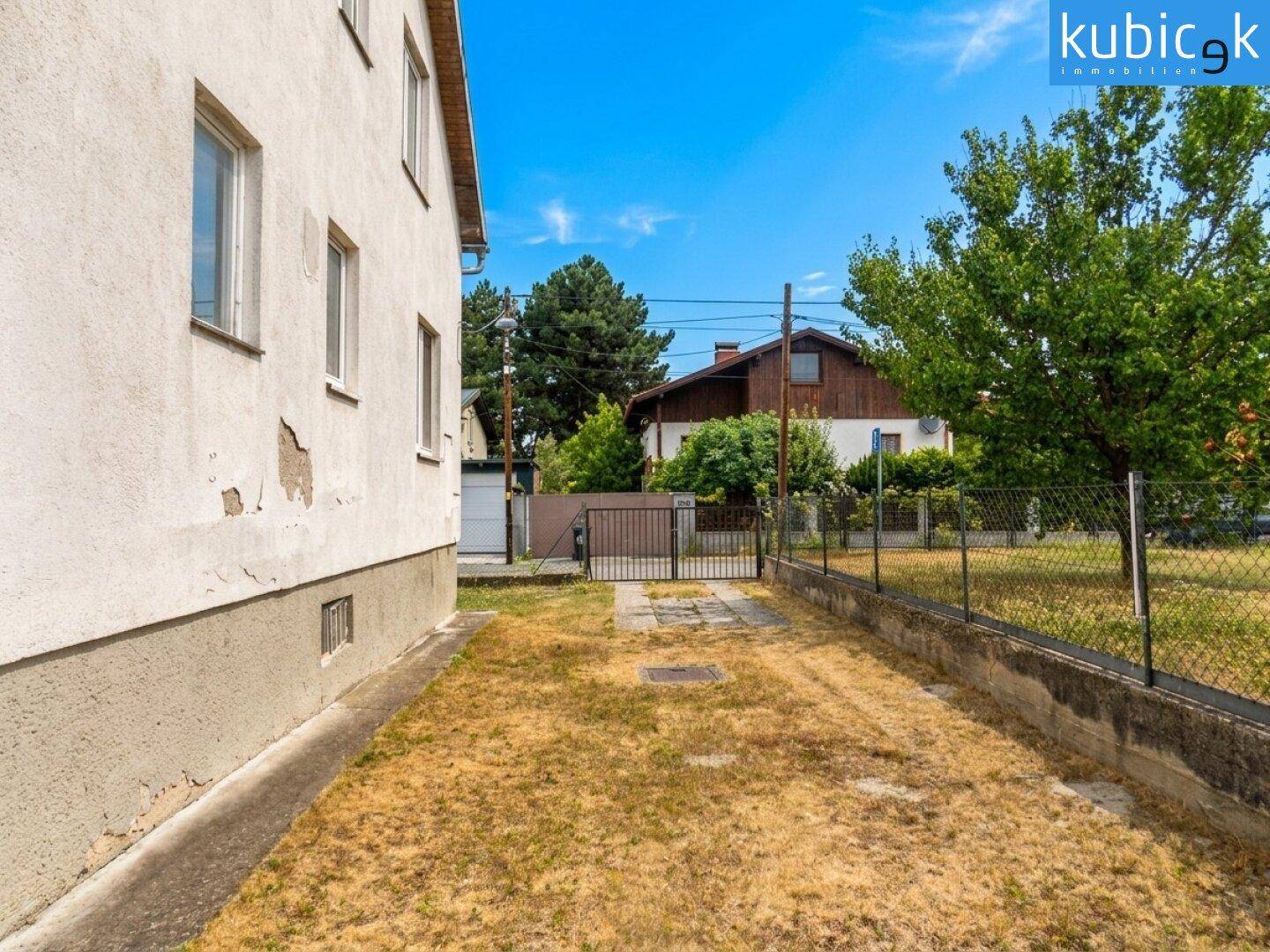 Seitenansicht des Hauses mit trockenem Rasen und einem Zaun unter blauem Himmel.