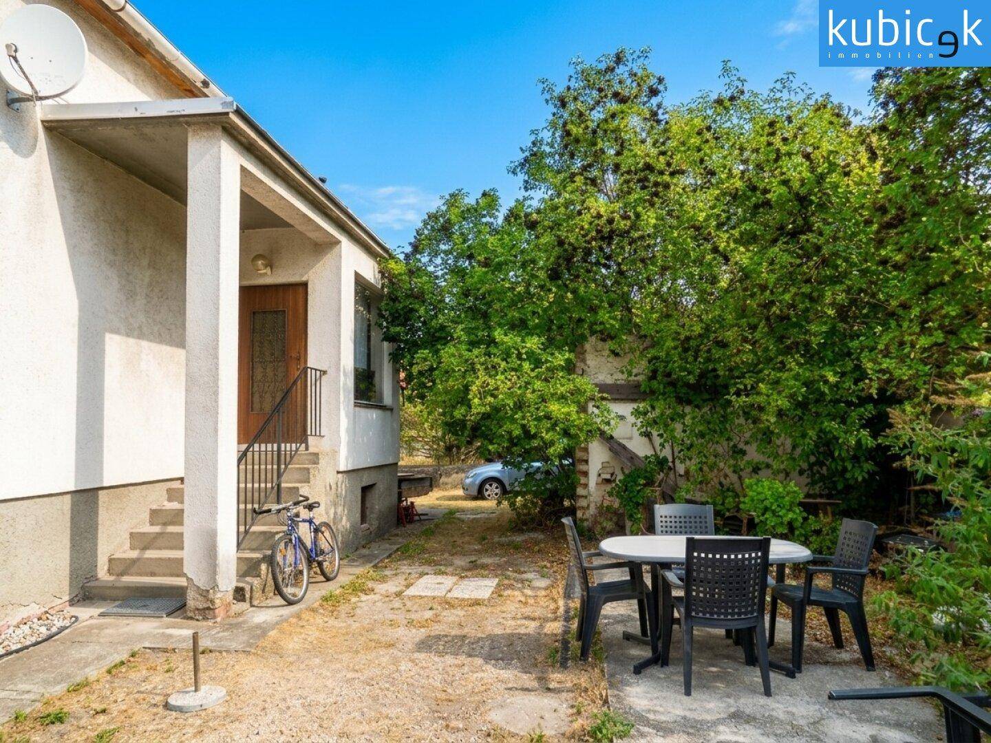 Eingangsbereich des Hauses mit einer kleinen Terrasse und Gartenmöbeln unter Bäumen.