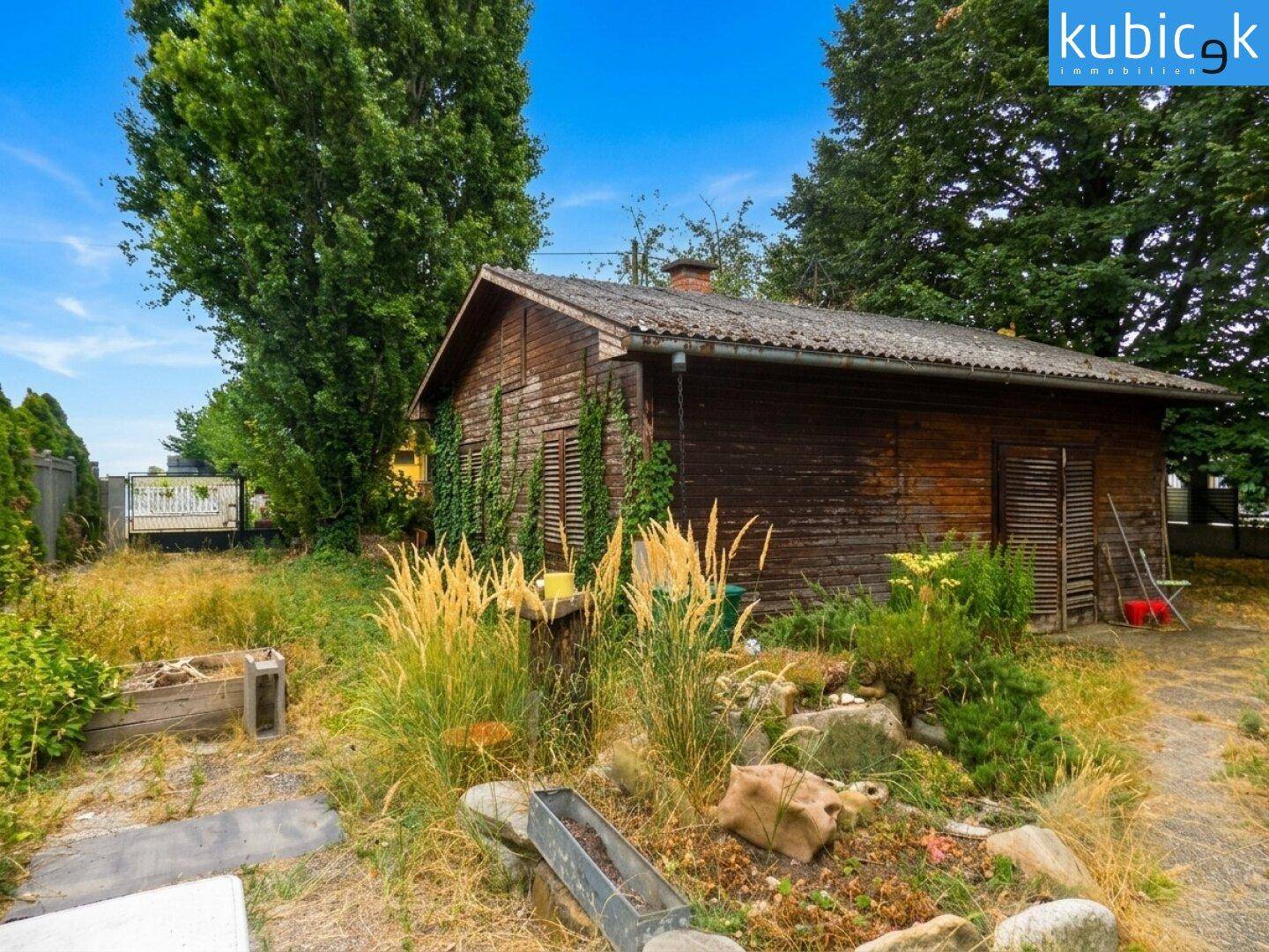 Rustikaler Holzschuppen im Garten mit umgebender Vegetation und Steinelementen.