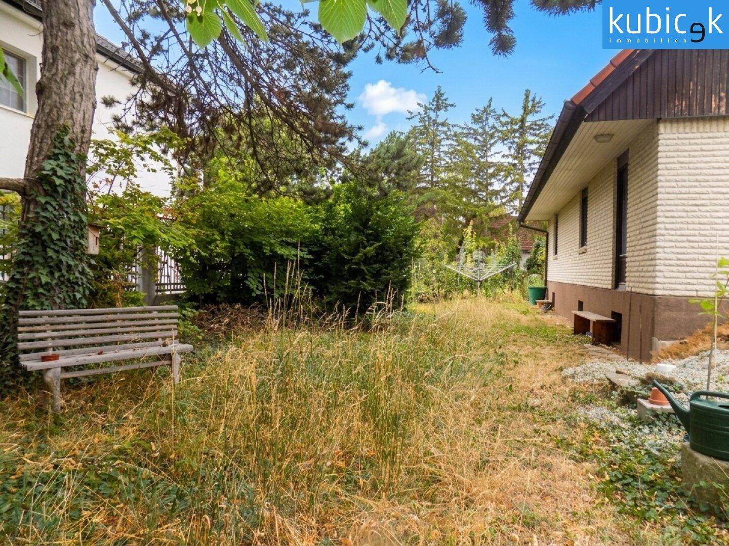 Verwilderter Gartenbereich mit alter Holzbank und hohen Gräsern neben dem Haus.