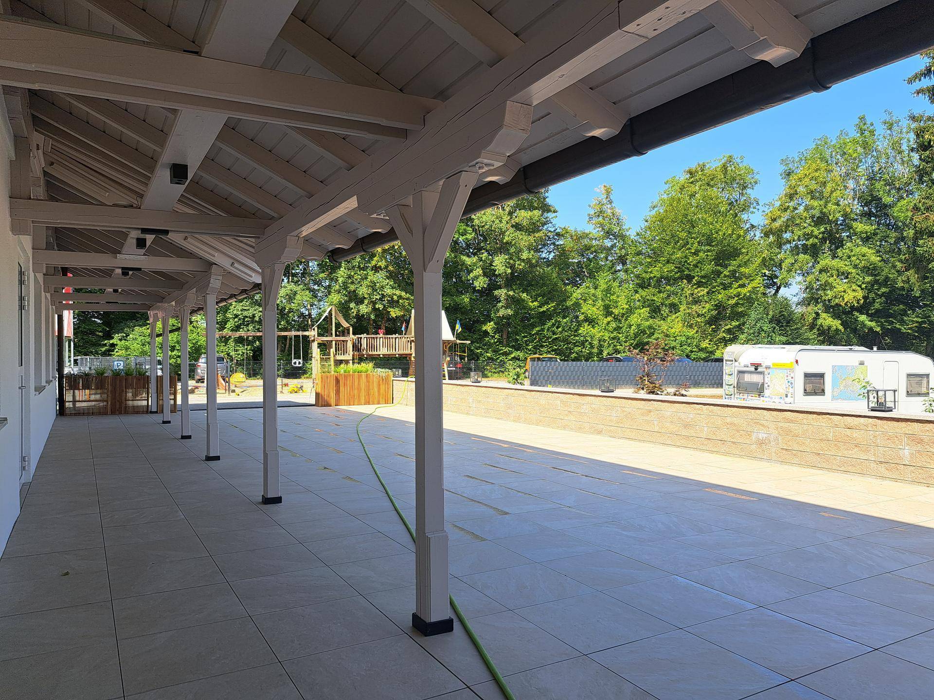 Überdachte Terrasse mit Holzbalken, die einen Blick auf Bäume und einen Spielplatz im Hintergrund bietet.