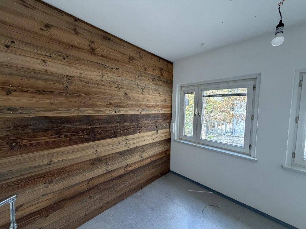 Raum mit einer markanten Holzwand, grauen Bodenfliesen und zwei Fenstern für natürliches Licht.