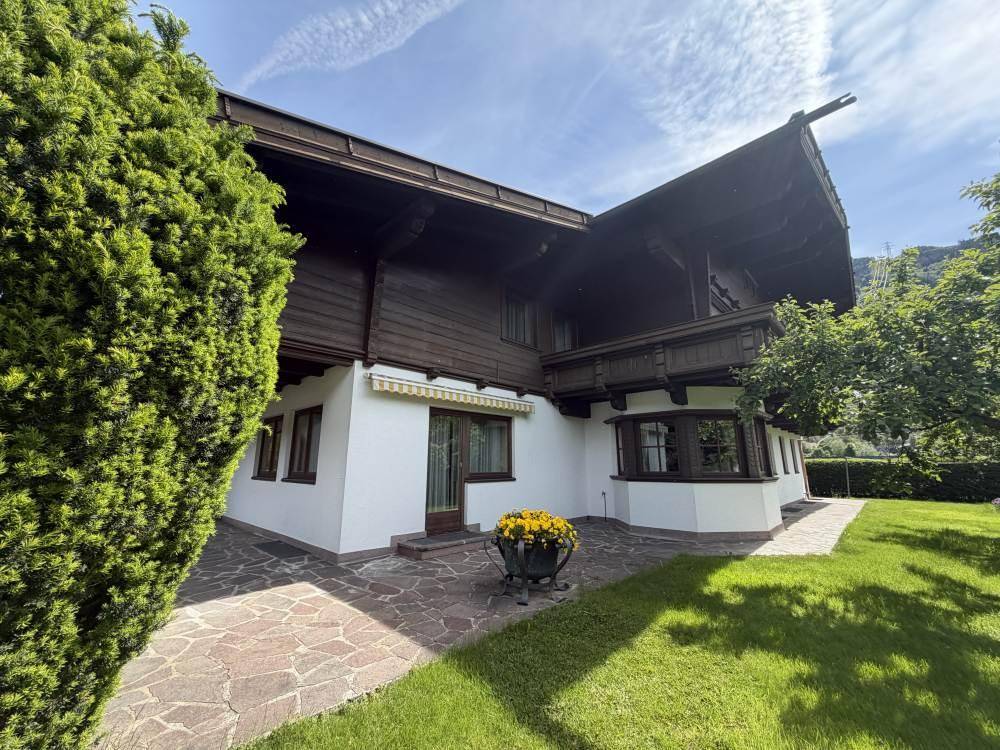 Hausfassade mit gepflasterter Terrasse, Rasen und üppiger Bepflanzung.
