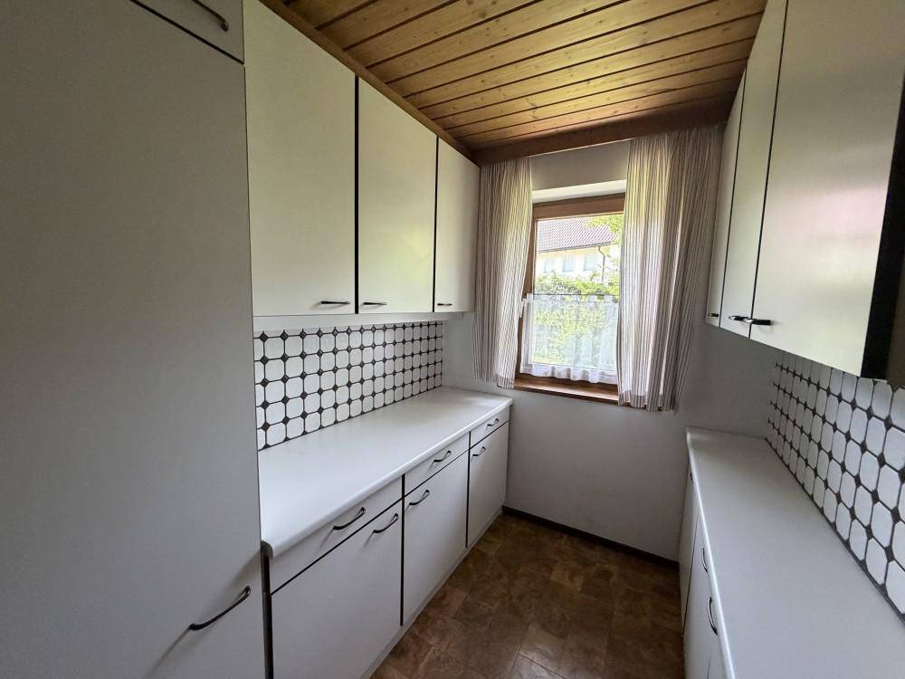 Schmale Küche mit weißen Schränken, gemusterten Fliesen und Fenster mit Gardine.