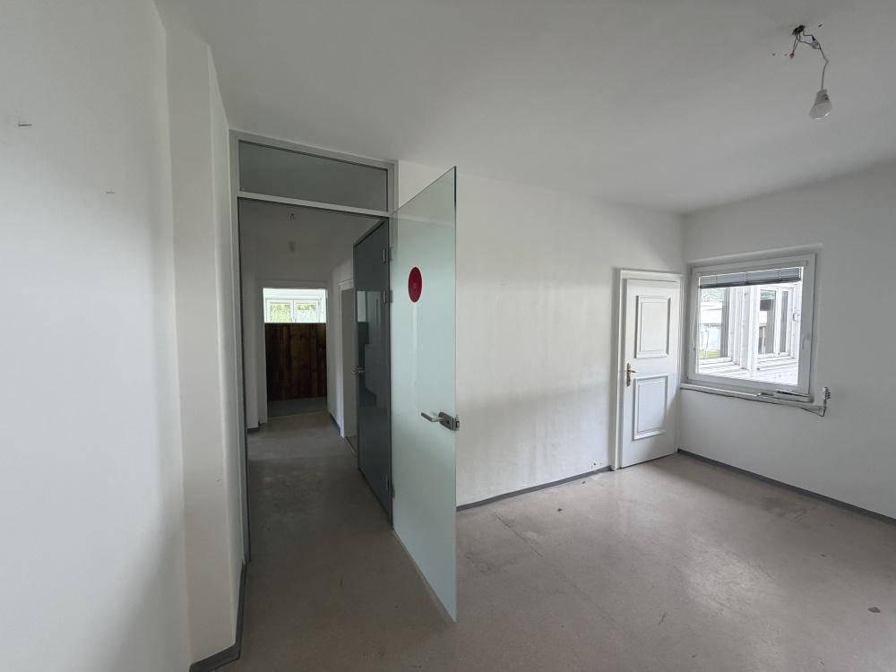 Leerer Raum mit grauen Bodenfliesen, einem Fenster und einer modernen Glastrennwand im Vordergrund.
