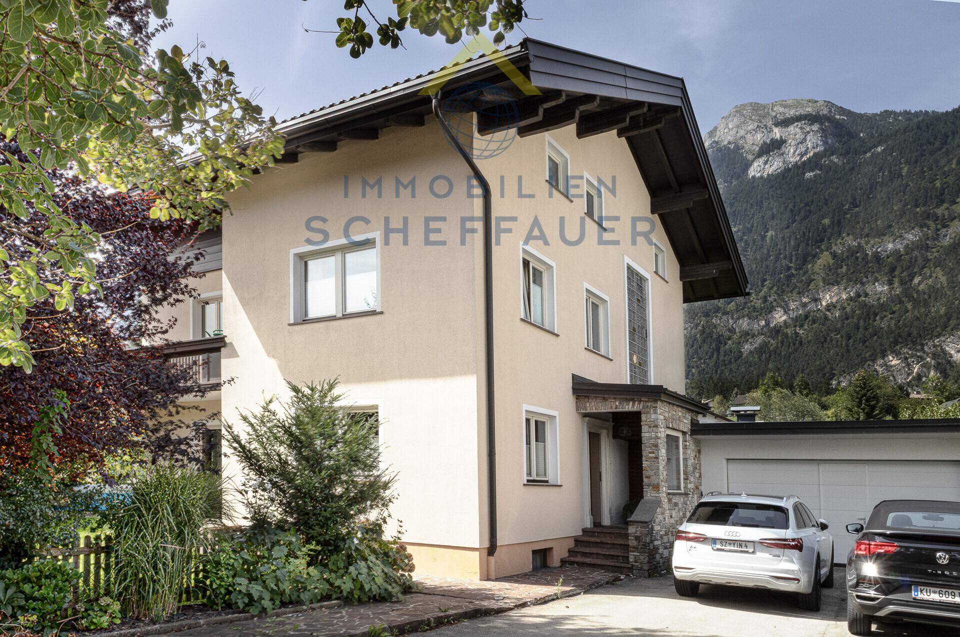 Vorderansicht des Hauses mit Einfahrt, Garage und gepflegter Fassade vor Bergkulisse.