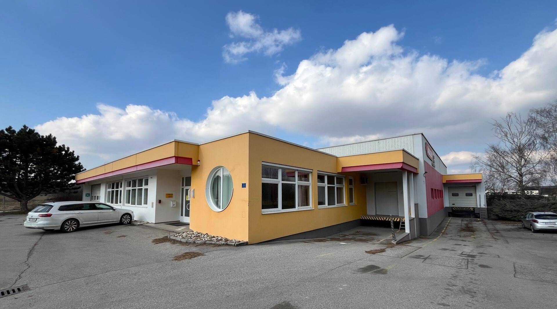 Gewerbegebäude mit gelber Fassade, großen Fenstern und einem überdachten Eingangsbereich unter bewölktem Himmel.