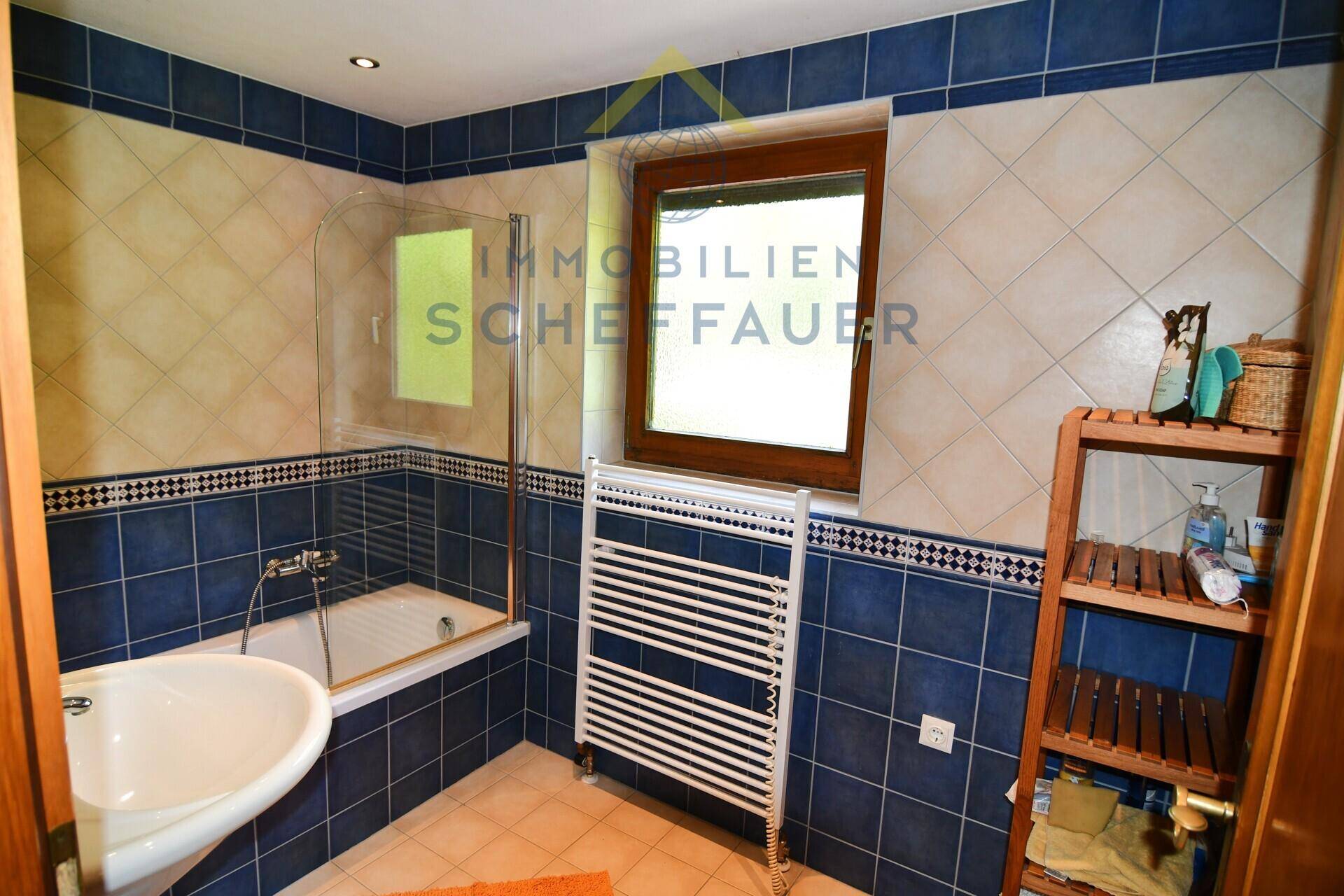 Badezimmer mit blauen und beigen Fliesen, Badewanne mit Duschabtrennung und Fenster.