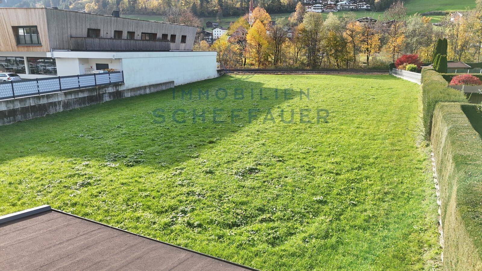 Blick über das grüne Grundstück mit Hecke und den dahinterliegenden Häusern und Hügeln.