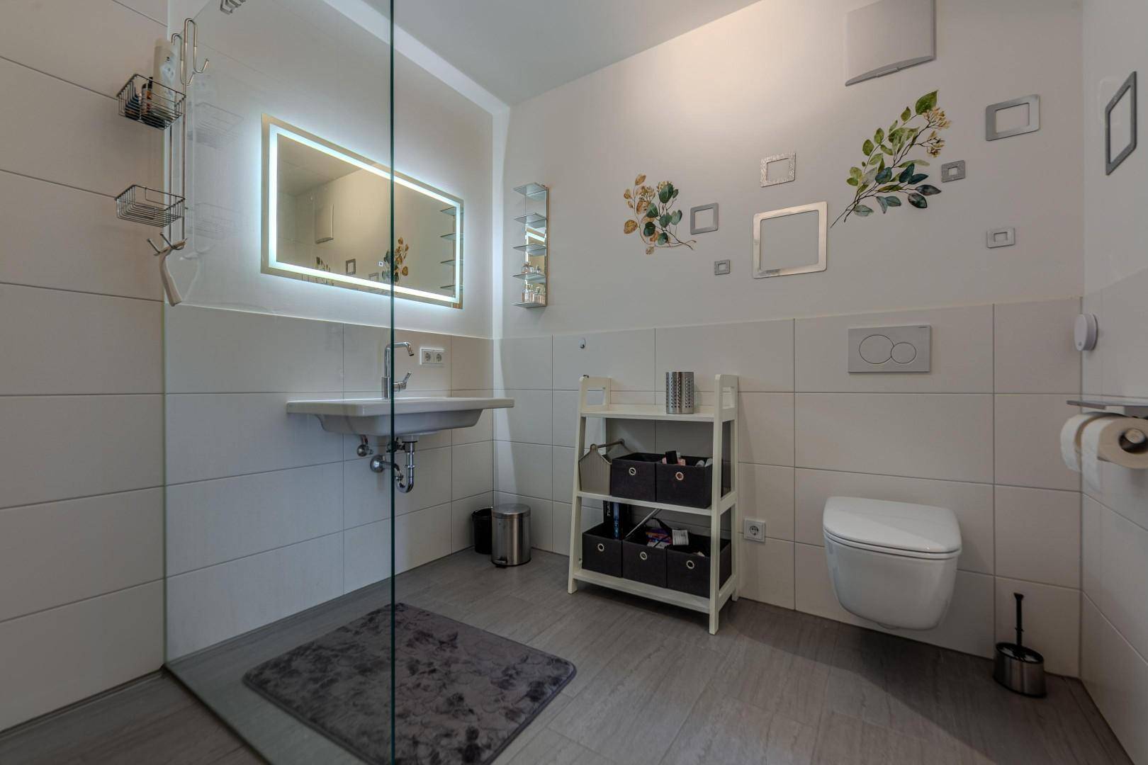 Modernes Badezimmer mit ebenerdiger Dusche, beleuchtetem Spiegel und Wand-WC.