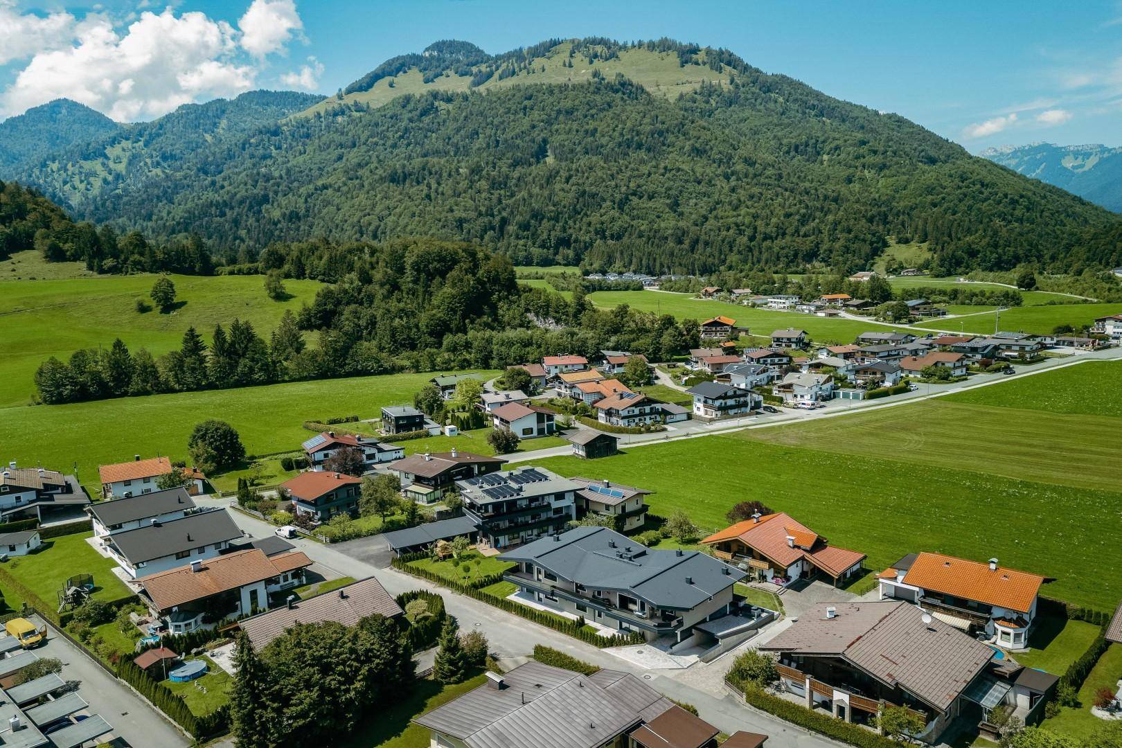 Luftaufnahme einer Wohnsiedlung mit Häusern und grünen Feldern vor einer beeindruckenden Bergkulisse.