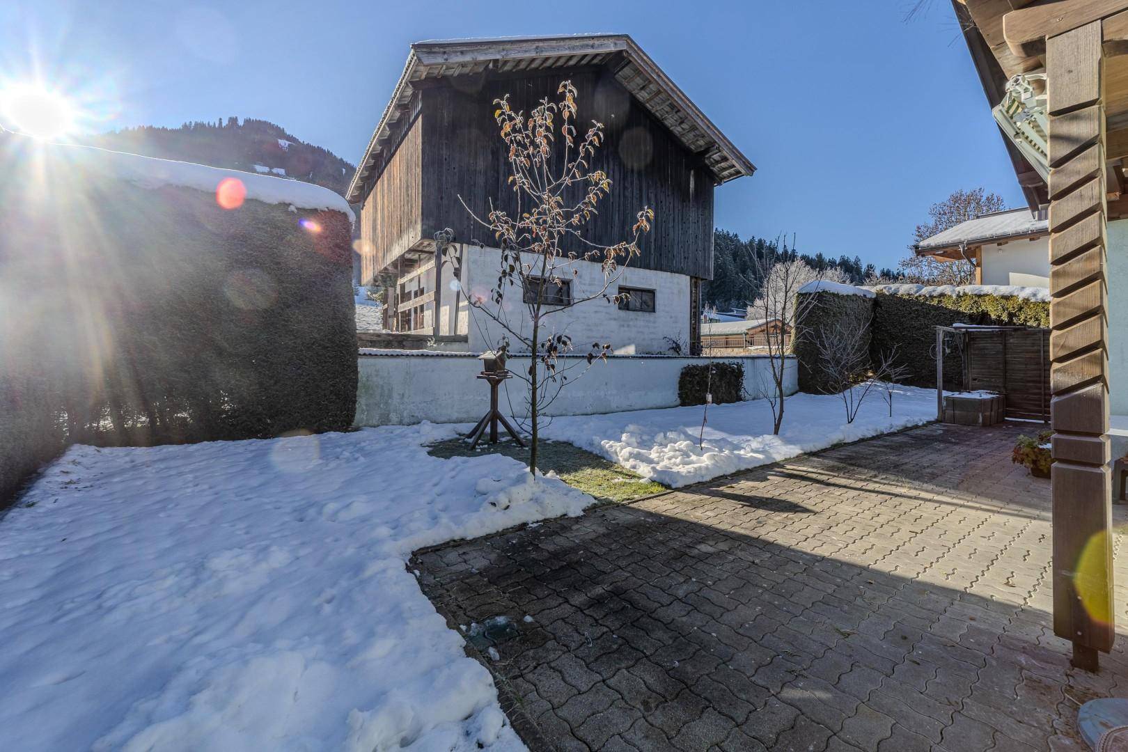 Rückansicht des Hauses mit verschneitem Garten und gepflastertem Bereich an einem sonnigen Wintertag.