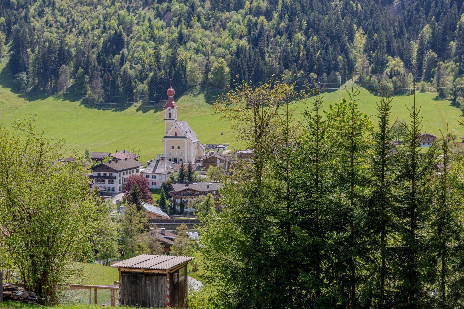 Panoramablick auf ein idyllisches Dorf mit einer Kirche, umgeben von grünen Hügeln und Wäldern.