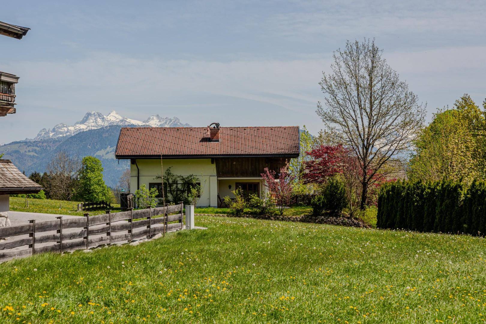 Traditionelles Haus mit rotem Ziegeldach, umgeben von grünen Wiesen und Bäumen mit Bergblick.