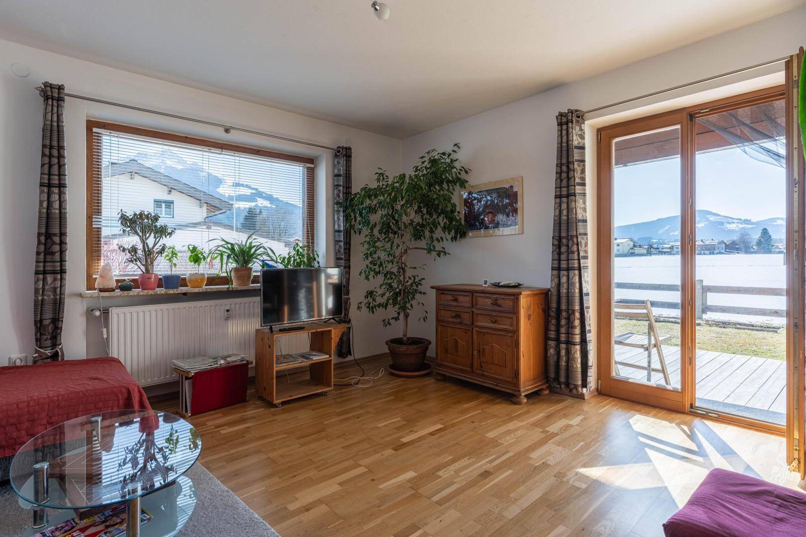 Gemütliches Wohnzimmer mit Holzboden, großen Fenstern und direktem Zugang zu einer Terrasse mit Bergblick.