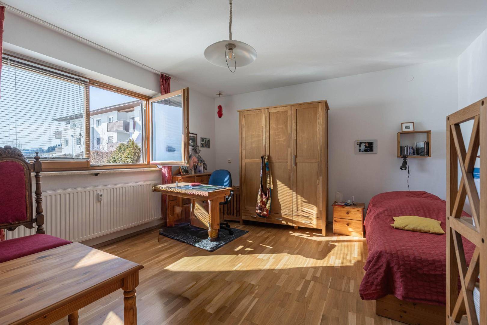 Helles Schlafzimmer mit Holzboden, Holzmöbeln und einem großen Fenster, das viel Tageslicht hereinlässt.