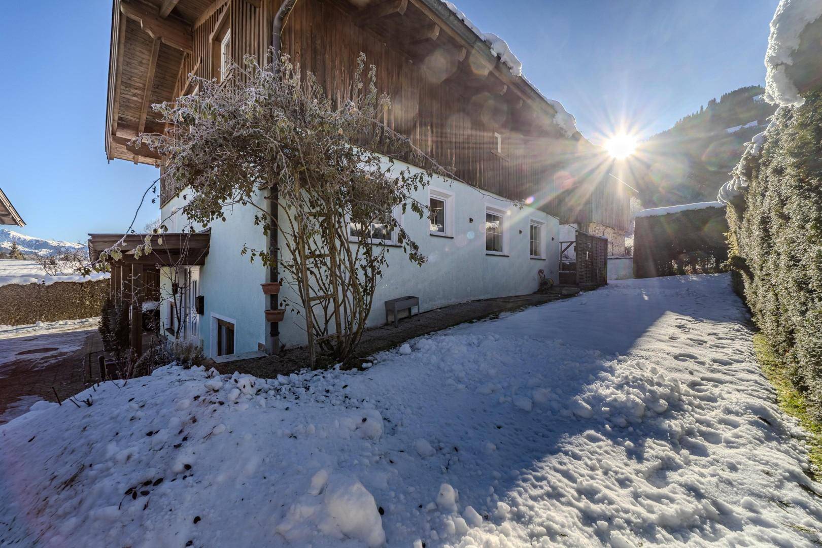 Hausfassade im Winter mit Schnee und Sonnenschein, umgeben von Bäumen.