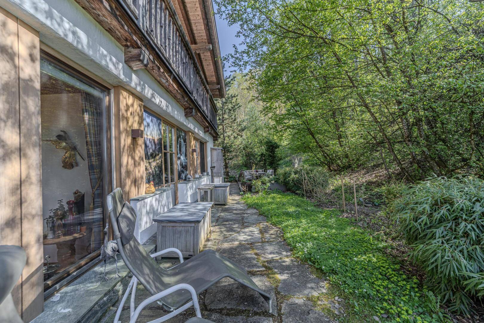 Seitlicher Außenbereich des Gebäudes mit Steinweg, Liegestuhl und dichter Bepflanzung.