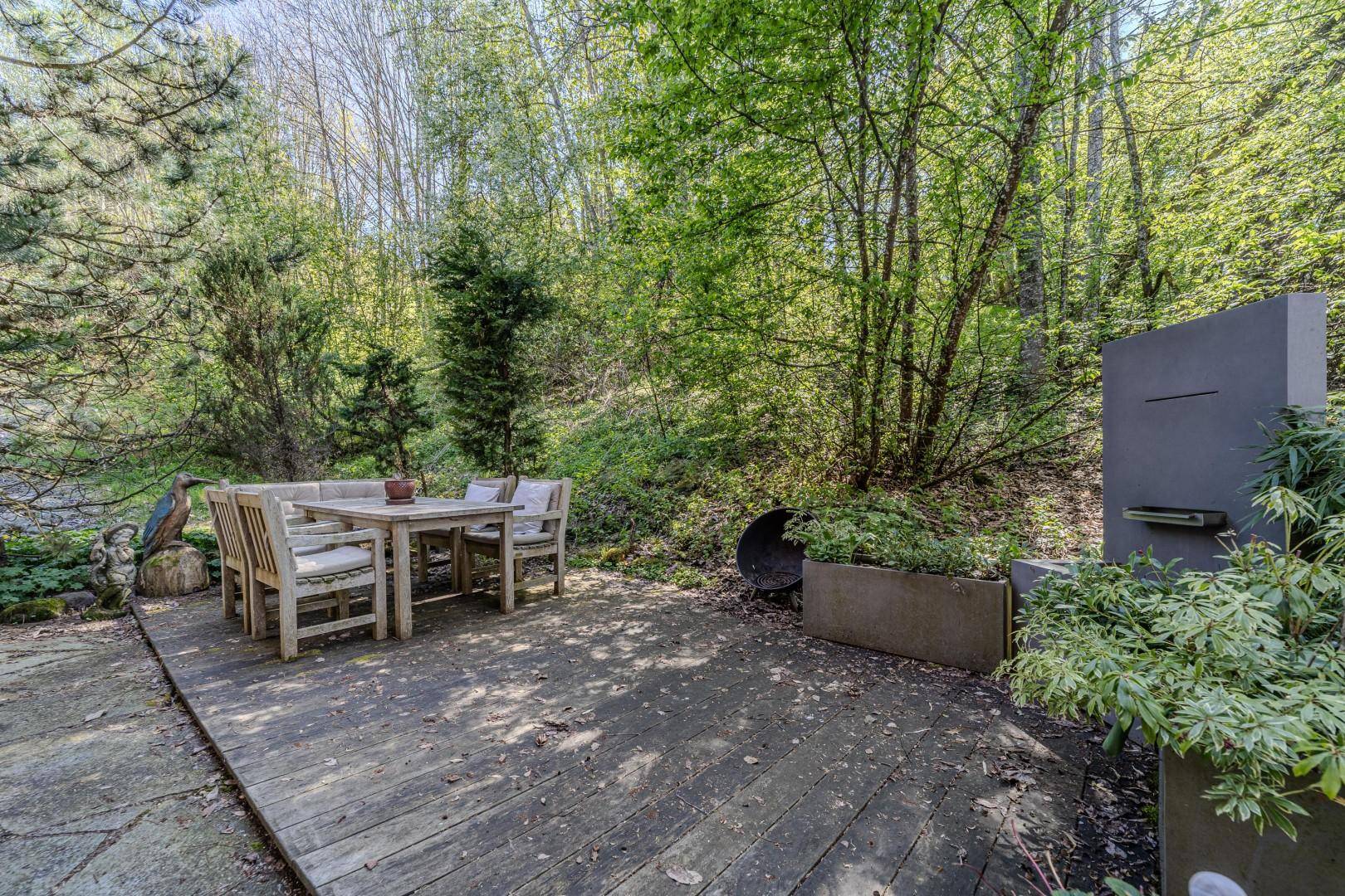 Holzterrasse mit Esstisch und Stühlen, umgeben von üppiger Vegetation und Bäumen.
