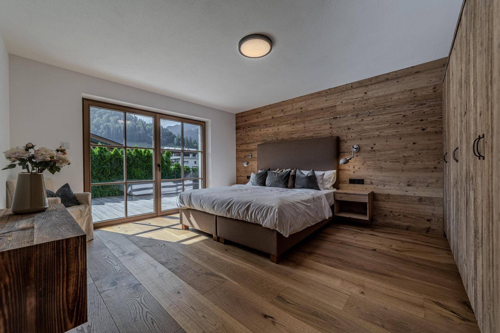 Geräumiges Schlafzimmer mit Holzwand, großen Fenstern und Blick ins Grüne.