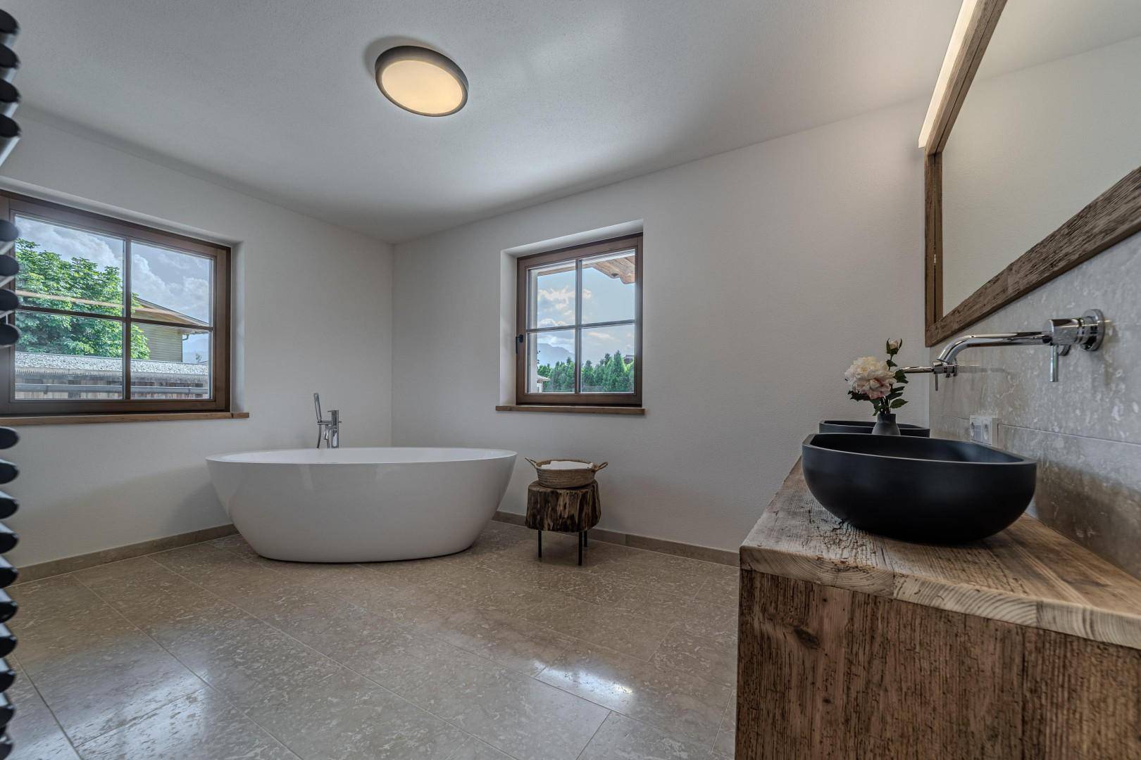 Geräumiges Badezimmer mit freistehender Badewanne, schwarzem Waschbecken und zwei Fenstern.