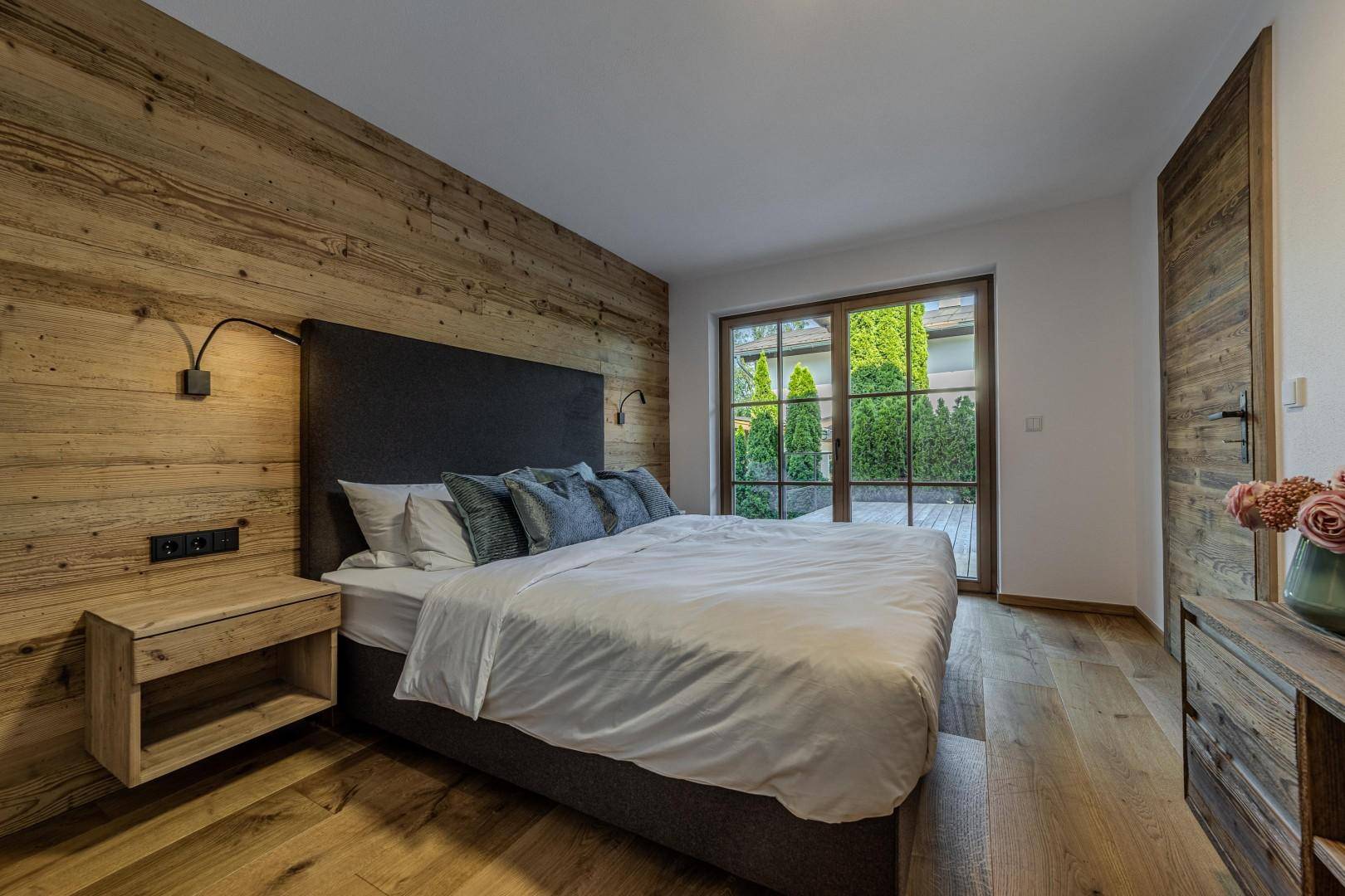 Helles Schlafzimmer mit Holzwand, großen Fenstern und Zugang zur Terrasse.
