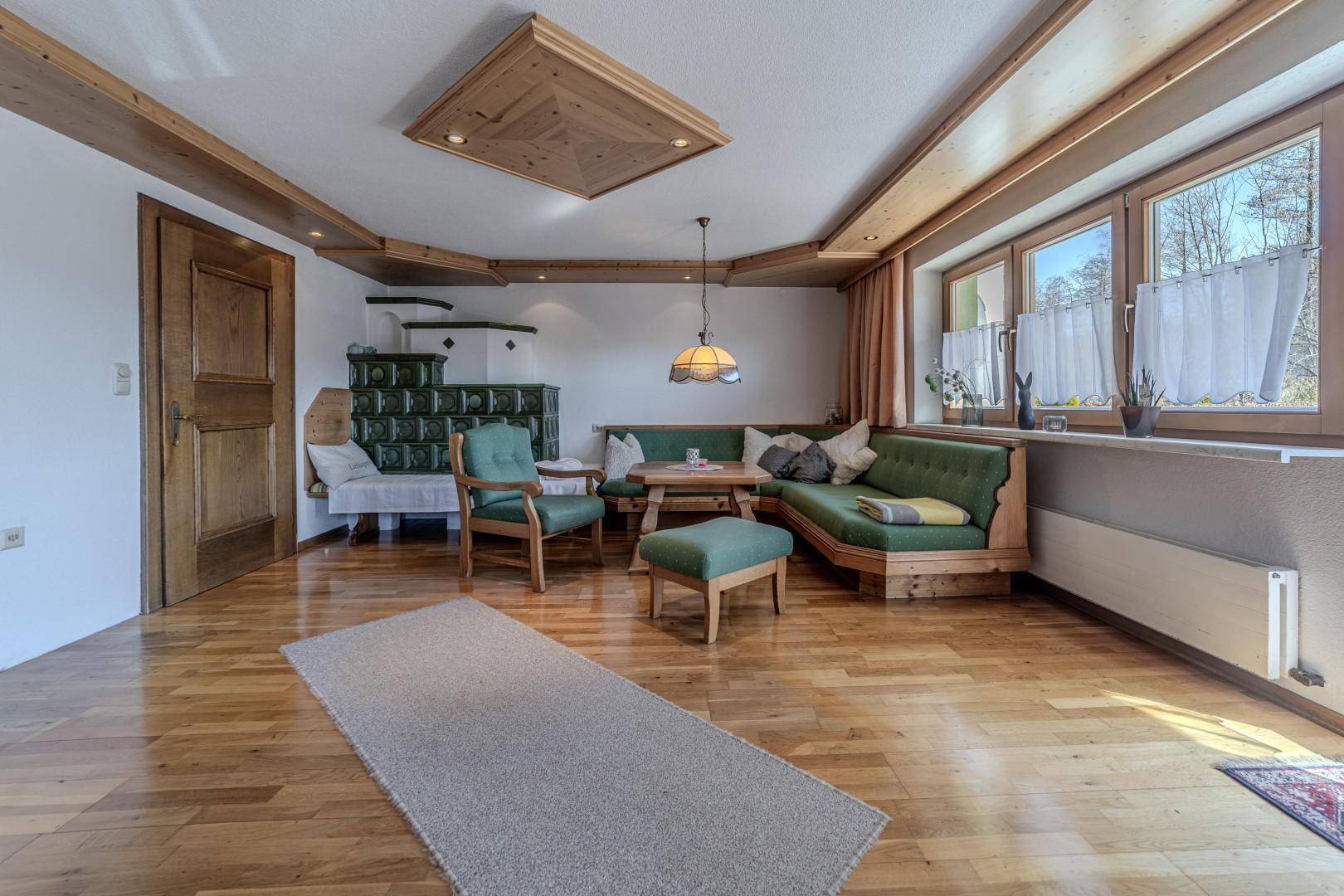 Helles Wohnzimmer mit großen Fenstern, Holzboden und einem traditionellen Kachelofen.