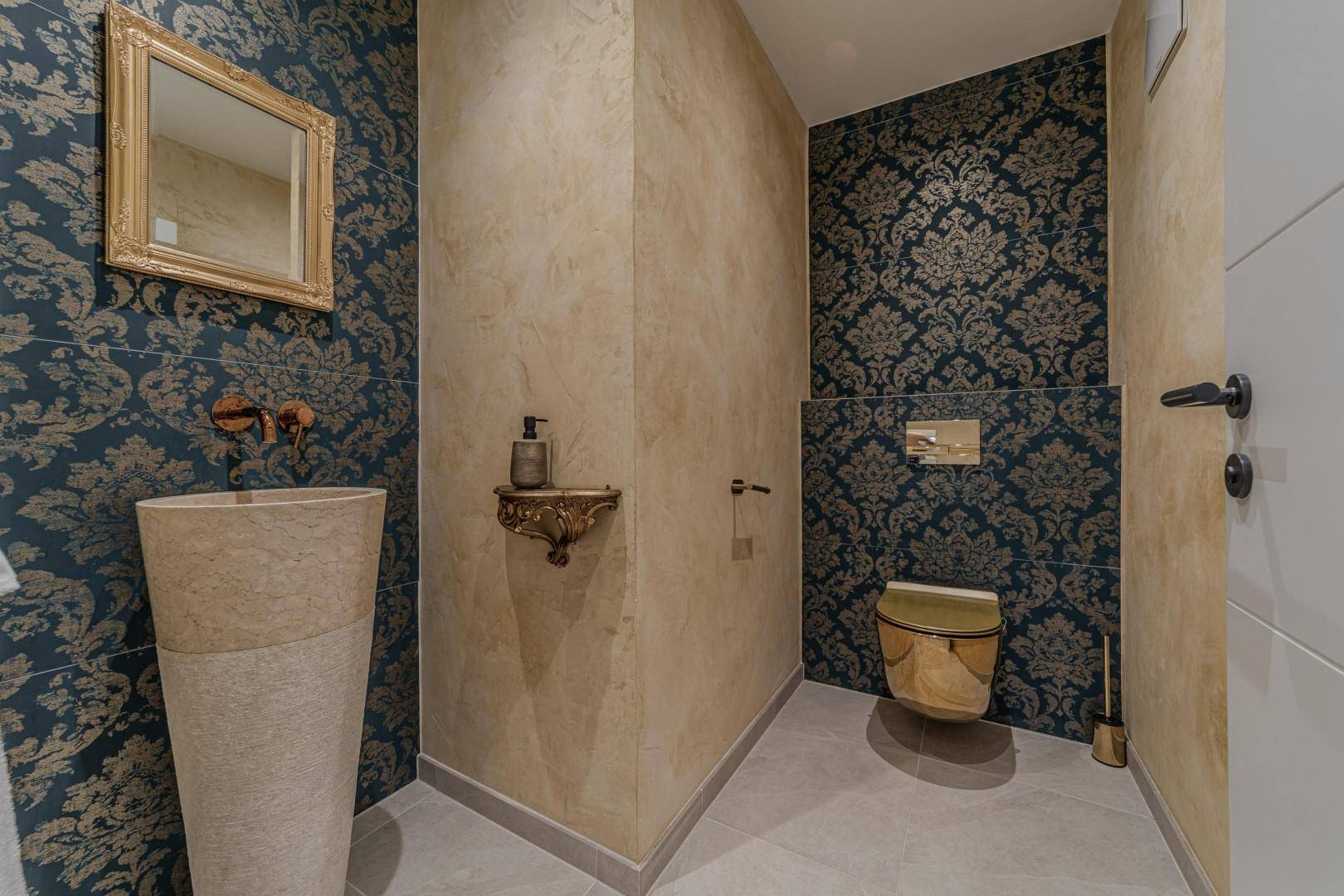 Exklusives Gäste-WC mit gemusterter Tapete, goldenem WC und stilvollem Steinsäulenwaschbecken.