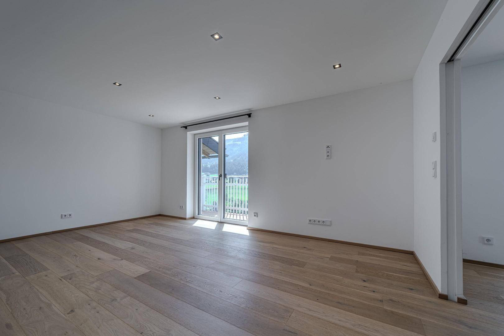 Leerer Raum mit Holzboden und weiß gestrichenen Wänden, mit Zugang zu einem sonnigen Balkon.