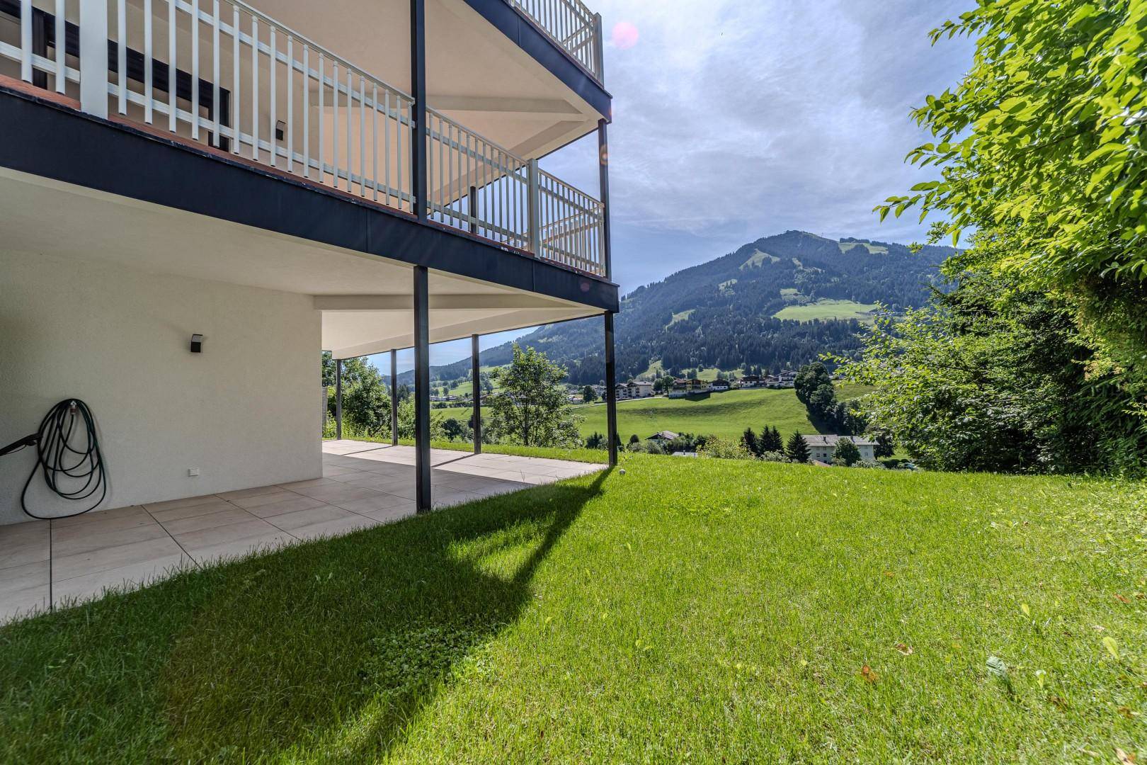 Sonniger Außenbereich mit Terrasse und Rasen, umgeben von grüner Natur und Bergen.