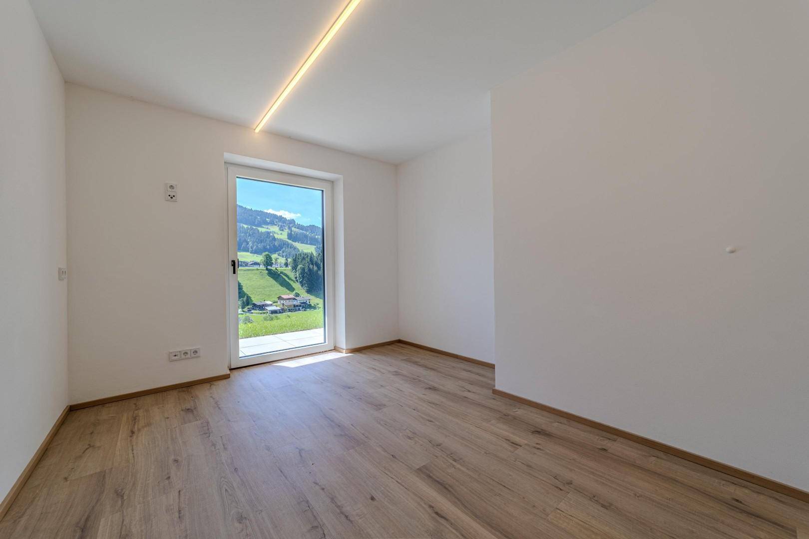 Heller Raum mit Holzboden, weißen Wänden und Zugang zu einer Terrasse mit Bergblick.