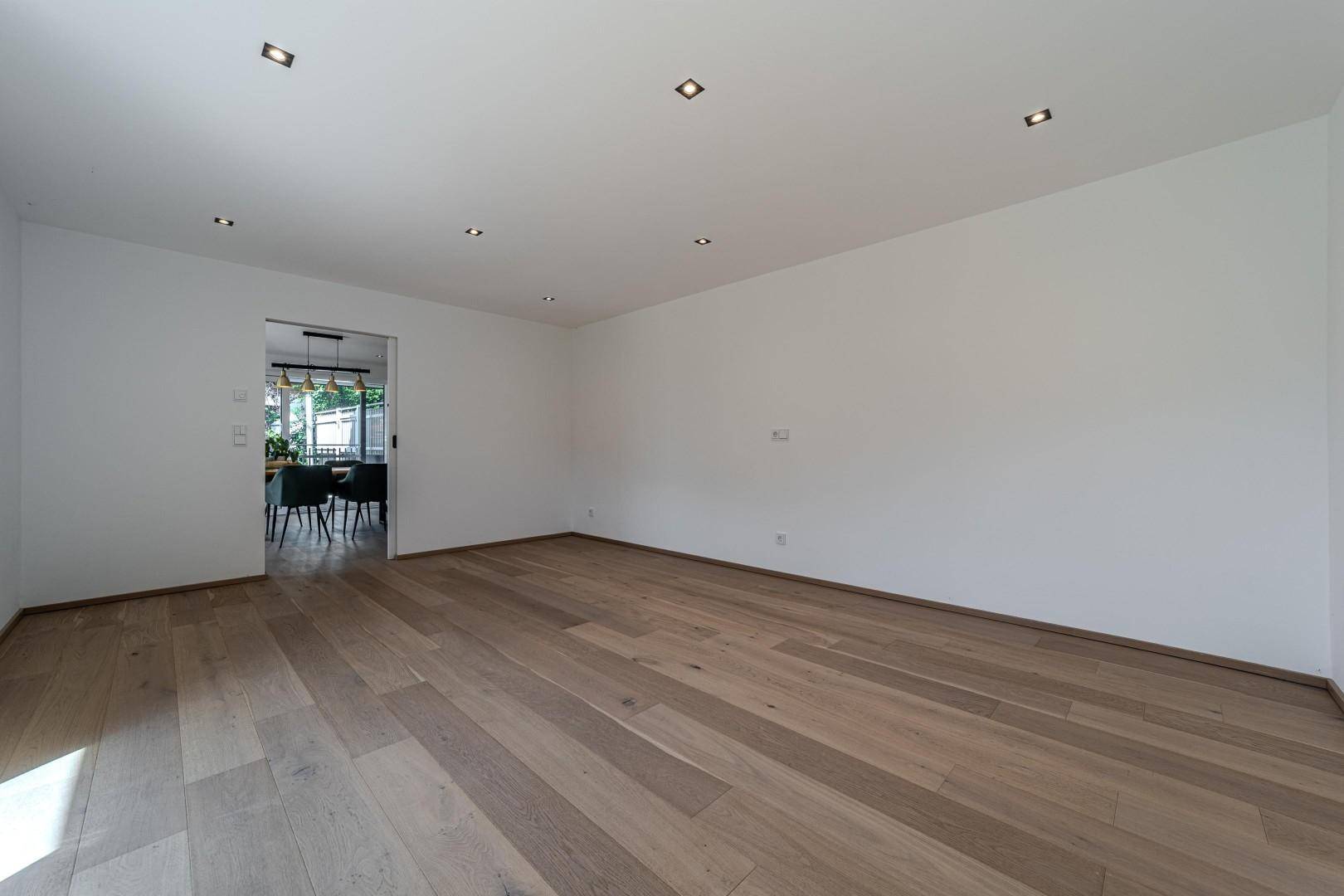 Geräumiger, leerer Raum mit Holzboden und offenem Zugang zu einem angrenzenden Essbereich.
