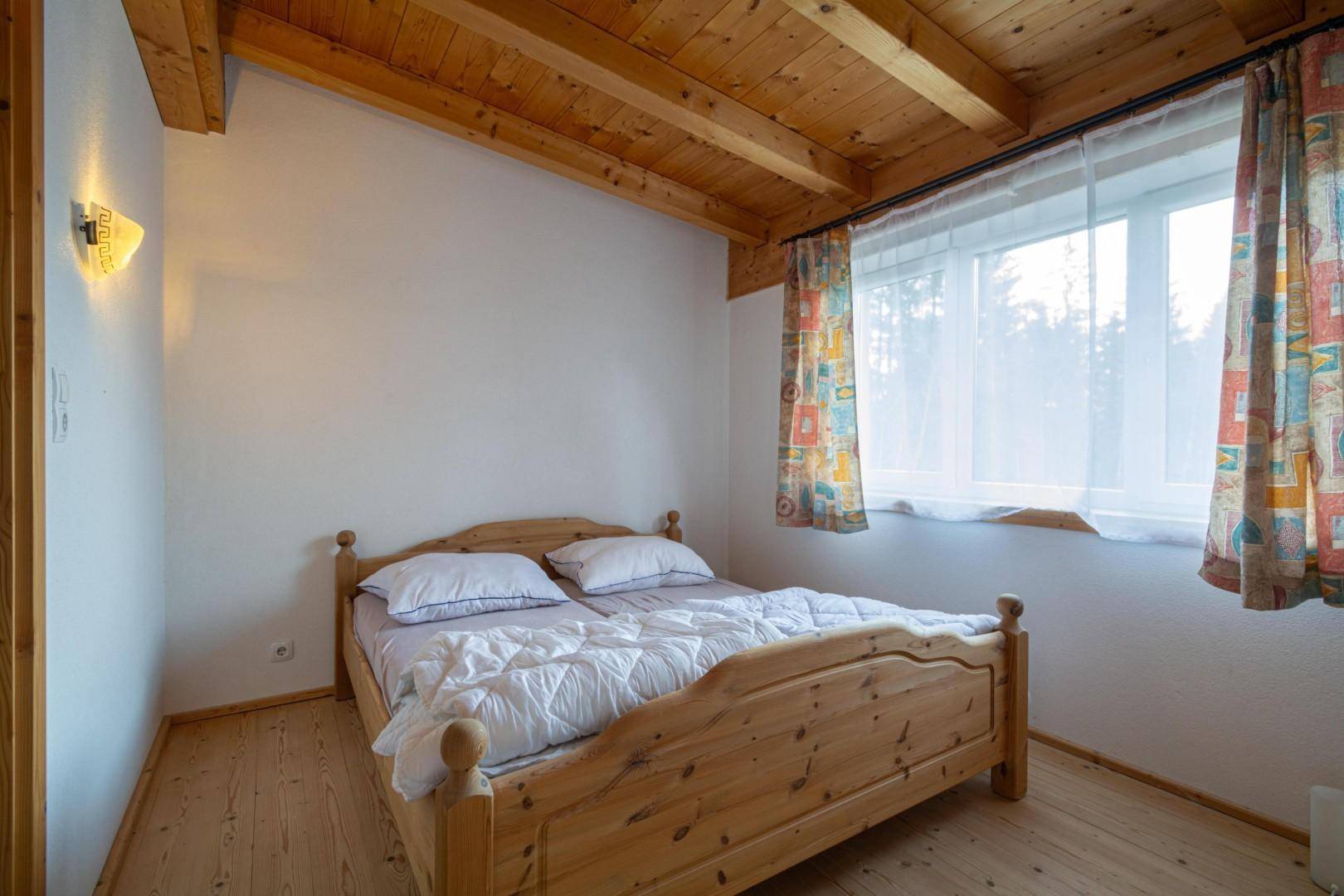 Einfaches Schlafzimmer mit Holzbett, hellen Wänden und einem Fenster mit bunten Vorhängen.