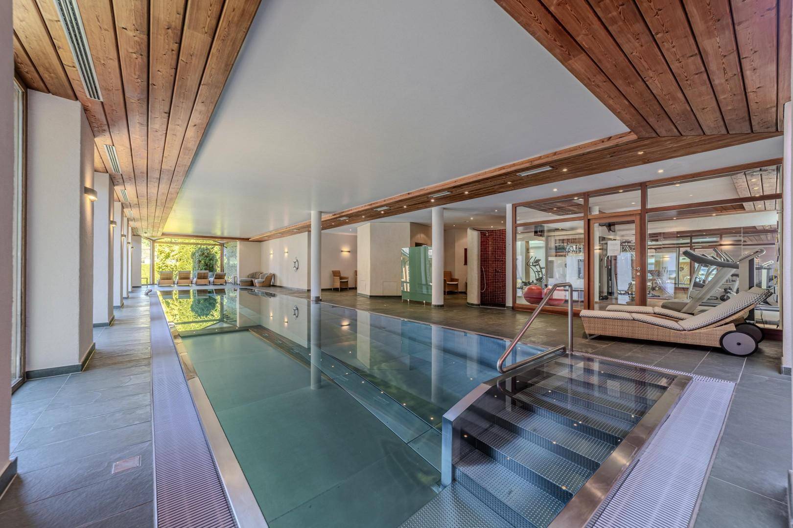 Großzügiger Innenpoolbereich mit Holzdecken, Liegestühlen und Blick auf den Fitnessraum.