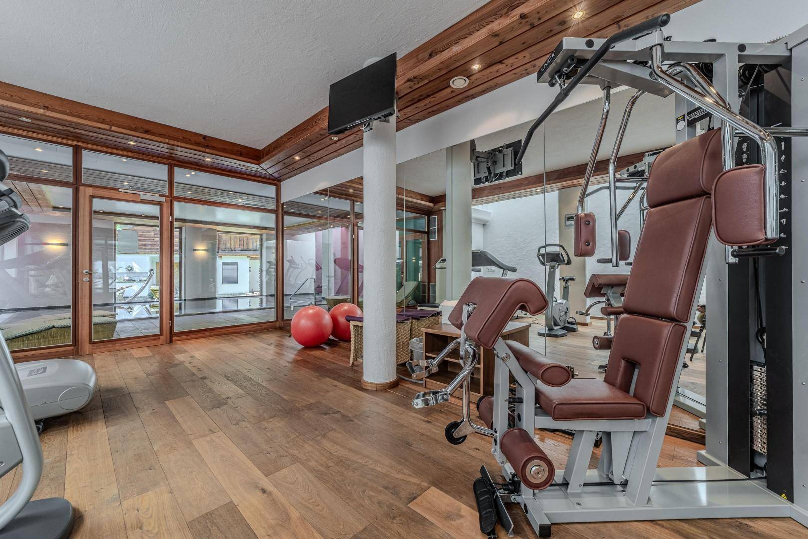 Moderner Fitnessraum mit Krafttrainingsgeräten, Holzboden und Blick auf den Poolbereich.