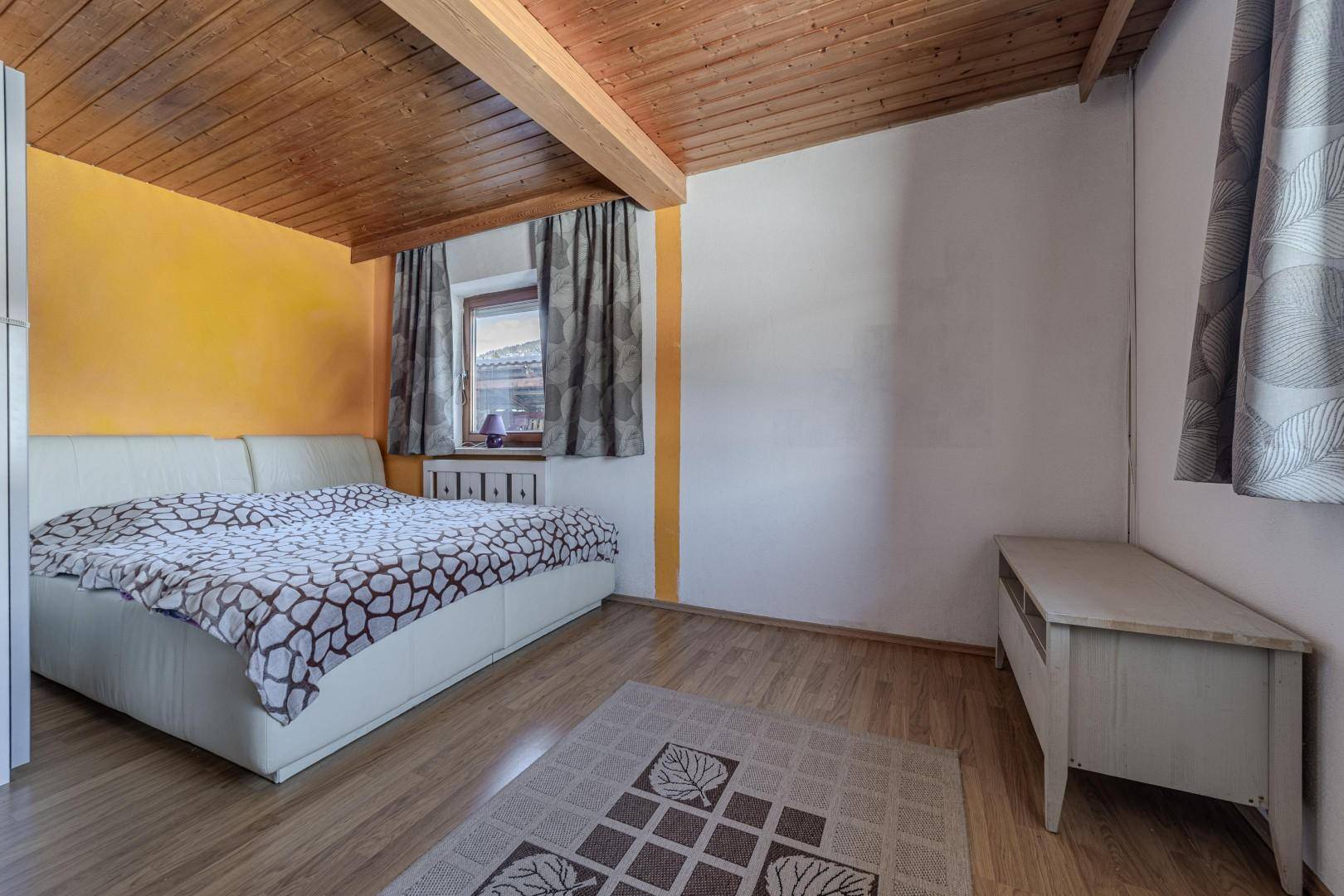 Schlafzimmer mit Holzbalkendecke, Holzboden, orangefarbener Akzentwand und Fenster.