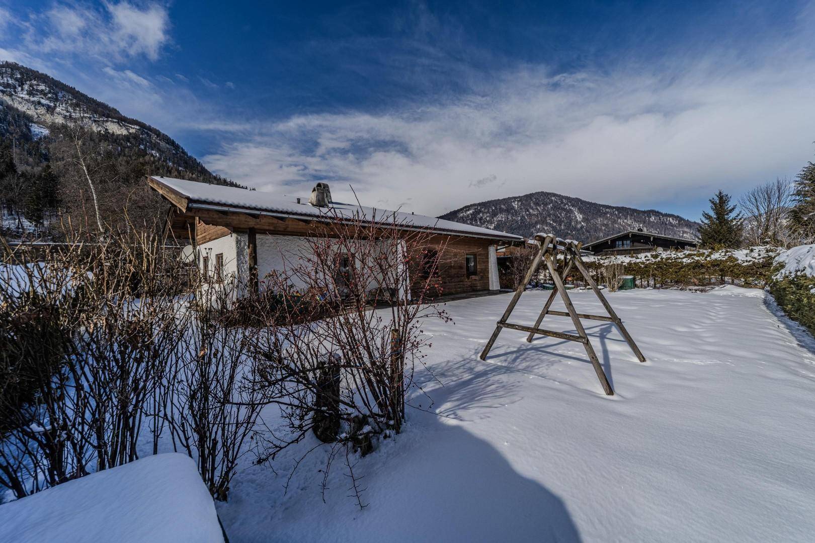 Hausansicht vom verschneiten Garten aus mit Schaukel und Bergpanorama.