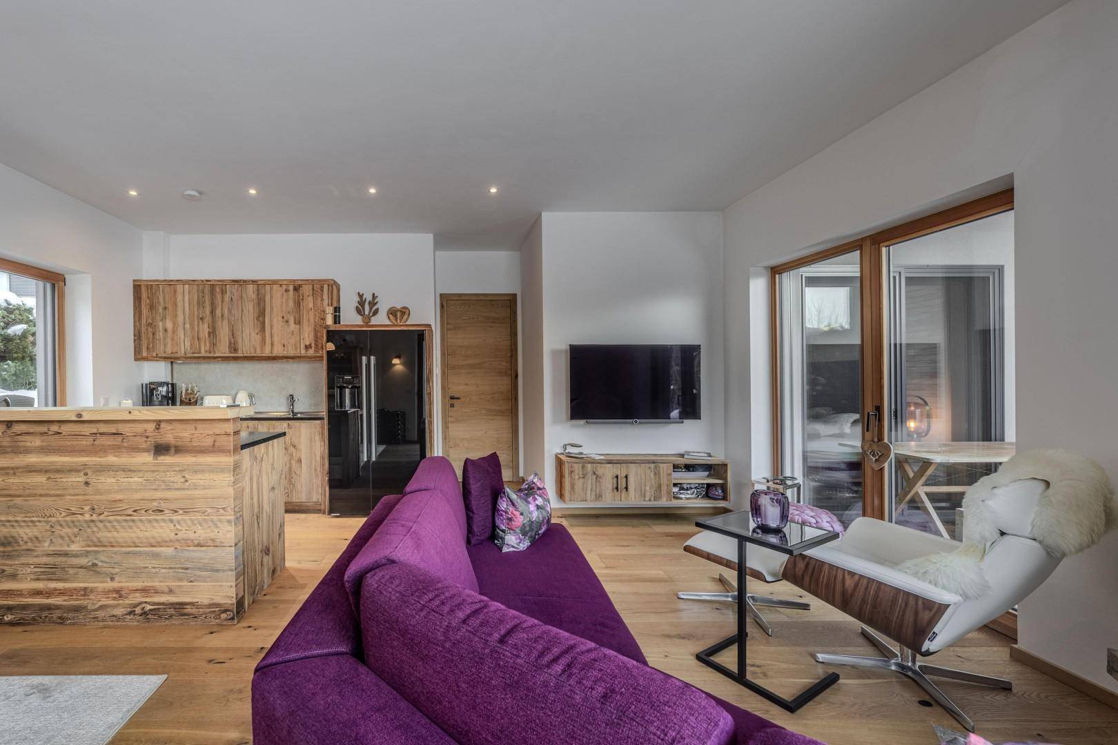Heller Wohnbereich mit bequemer Couch, Fernseher und offener Küche mit Holzelementen.