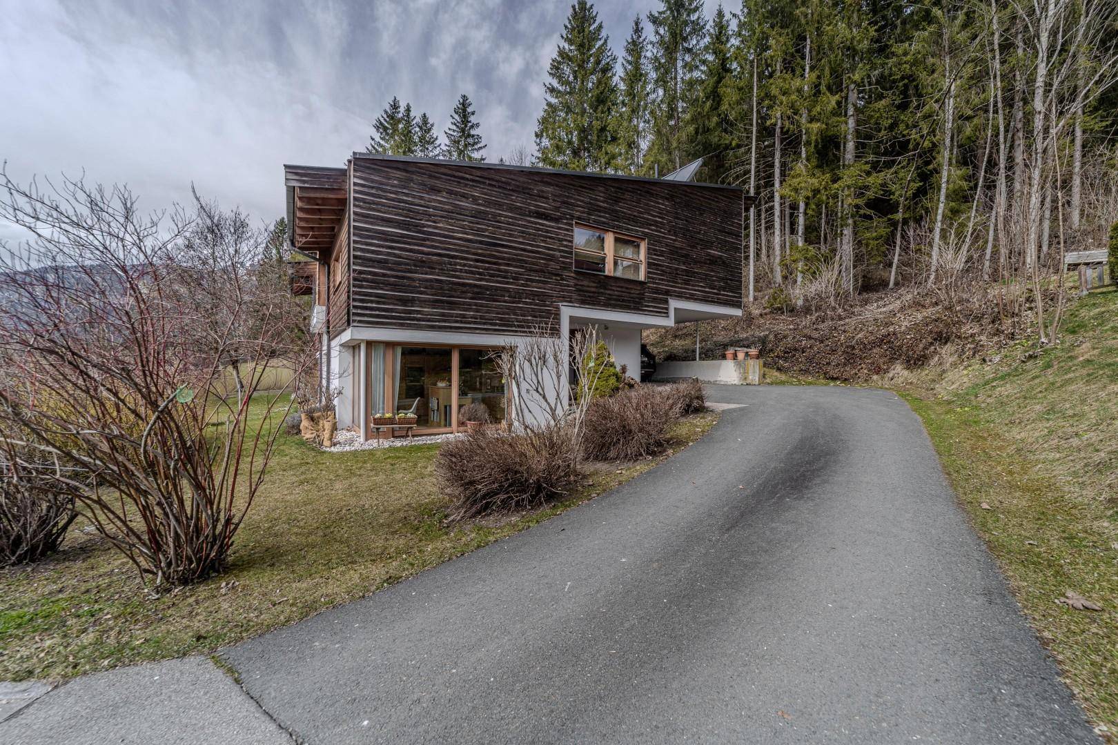 Seitenansicht des Hauses mit Zufahrt, umgeben von Bäumen und natürlicher Landschaft.