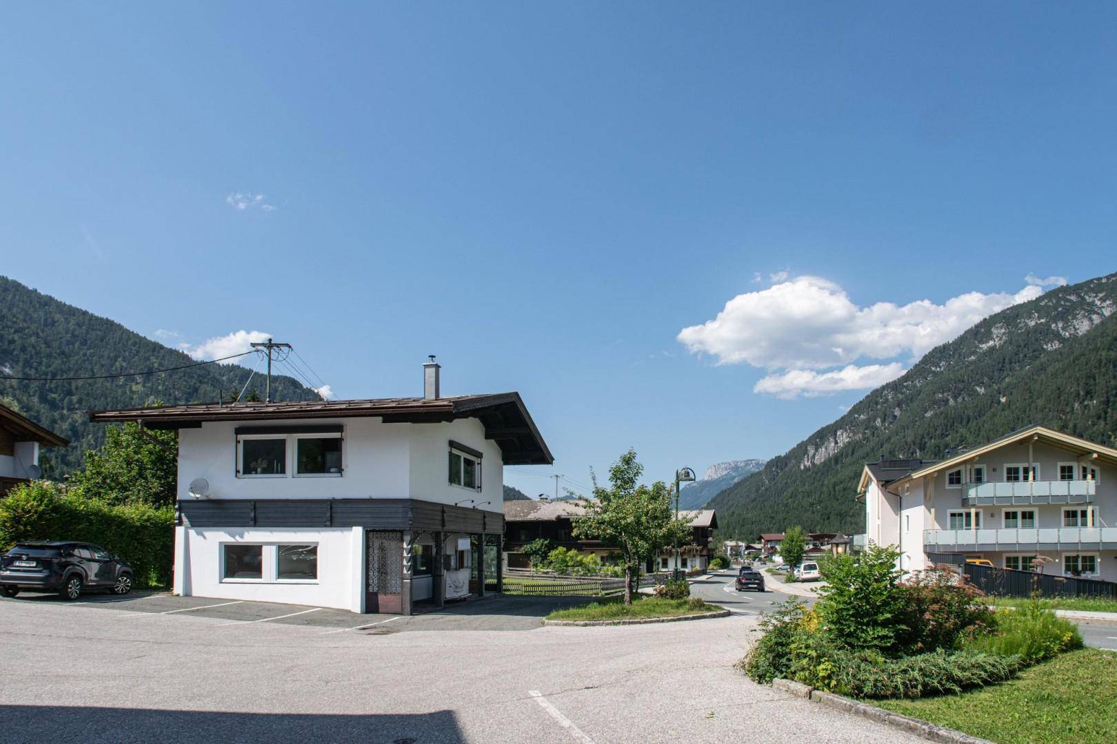 Ein weißes Gebäude mit dunklem Dach und Parkplätzen, umgeben von Bergen unter blauem Himmel.