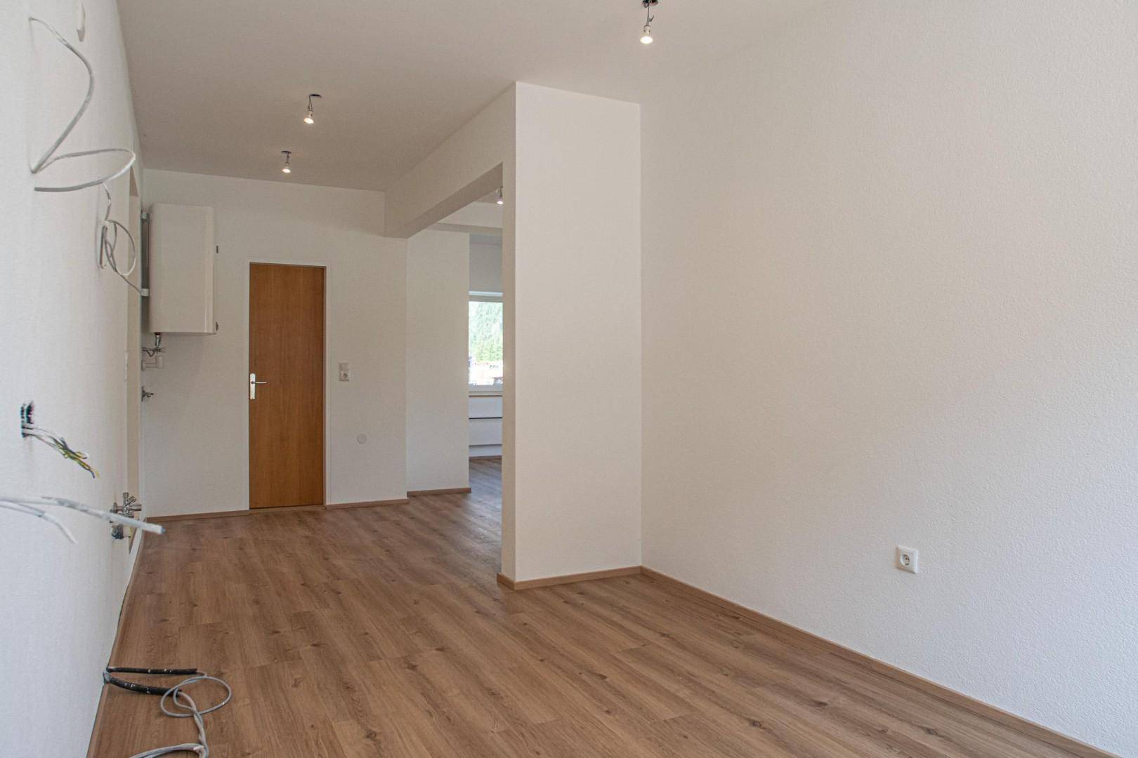 Offener Wohnbereich mit Laminatboden, weiß gestrichenen Wänden und einer Holztür.