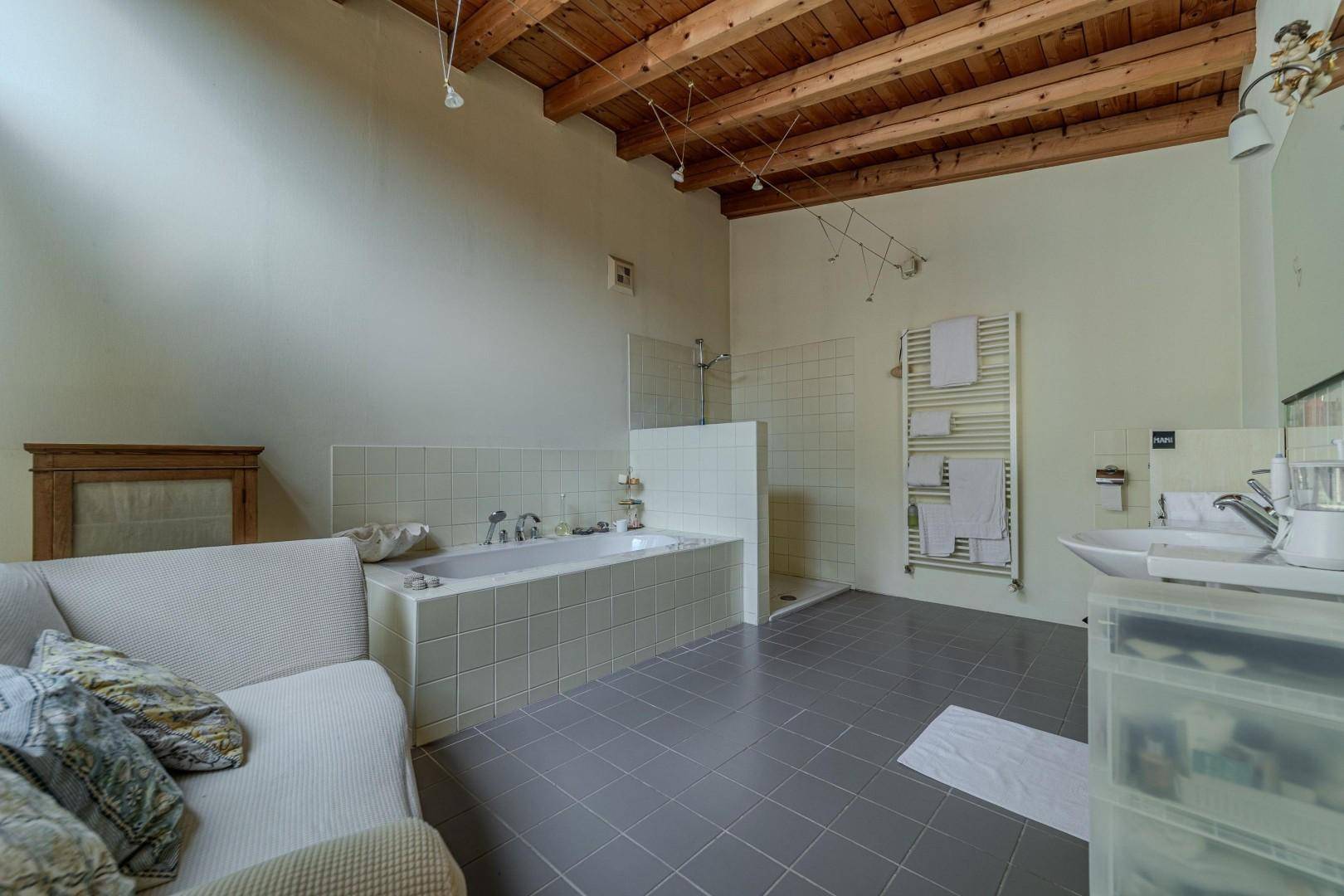 Großes Badezimmer mit Badewanne, Dusche, Fliesenboden und Holzbalkendecke.