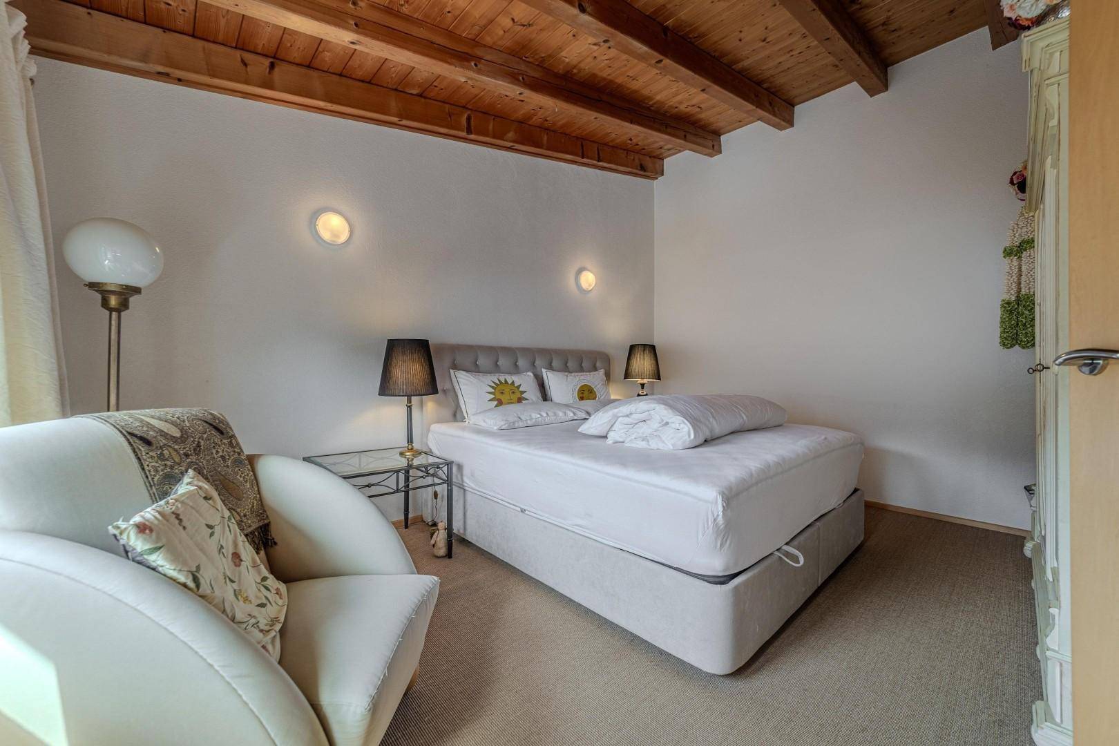 Helles Schlafzimmer mit Holzbalkendecke, Doppelbett und gemütlichem Sessel.