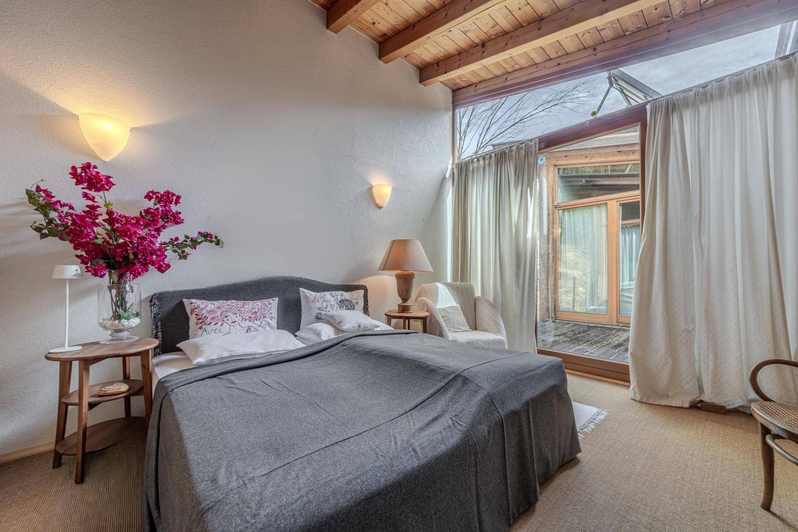 Schlafzimmer mit Holzbalkendecke, großem Fenster und Zugang zu einem Außenbereich.