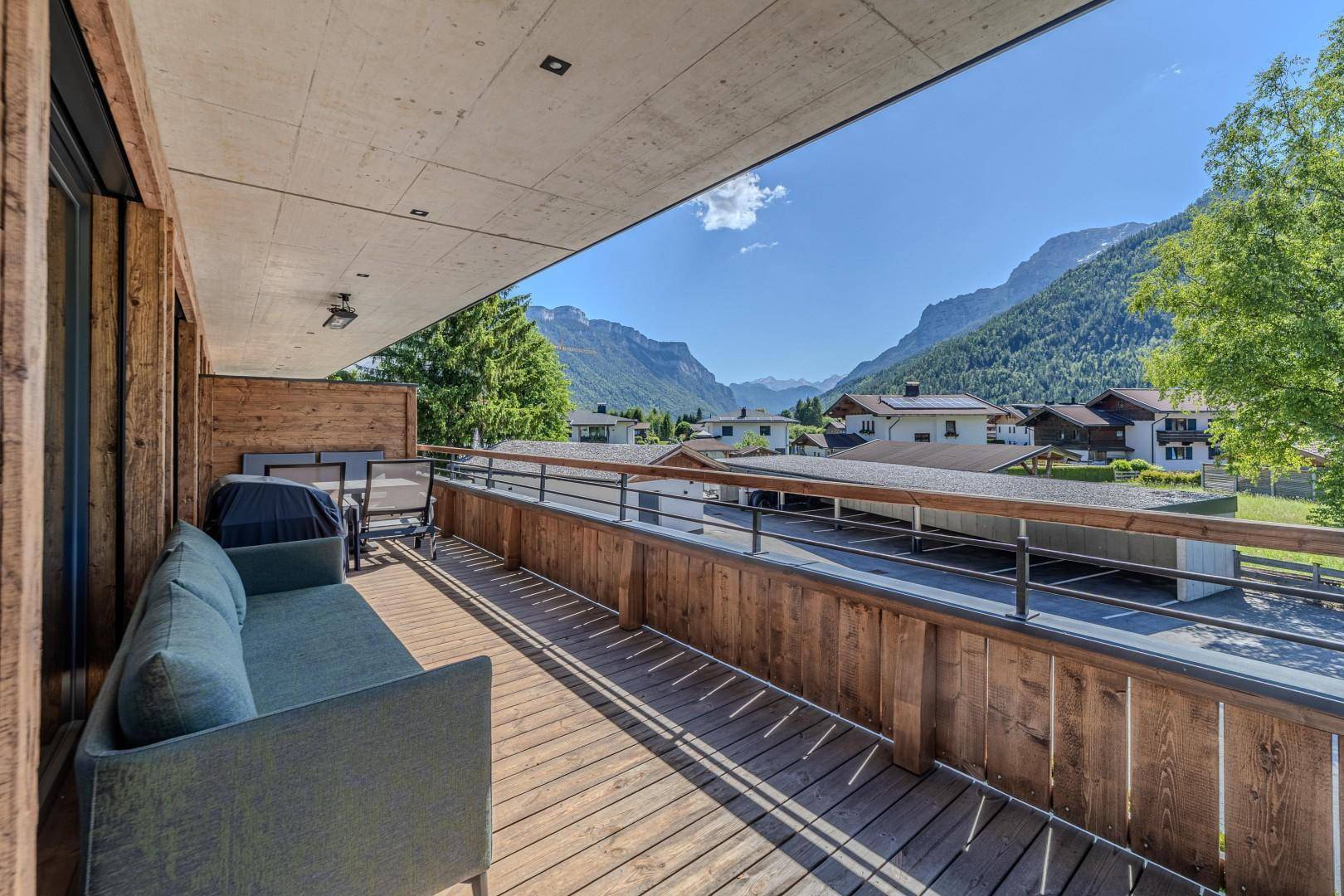 Sonniger Holzbalkon mit Lounge-Sofa und Panoramablick auf die Berge und das Tal.