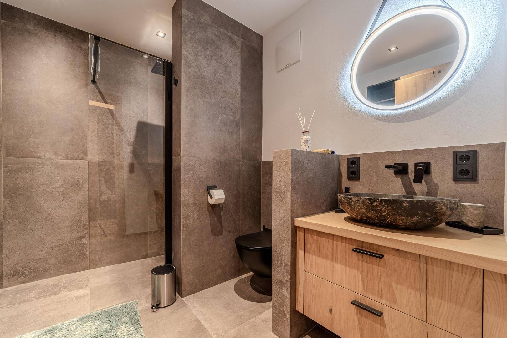 Modernes Badezimmer mit begehbarer Dusche, beleuchtetem Spiegel und dunklen Fliesen.