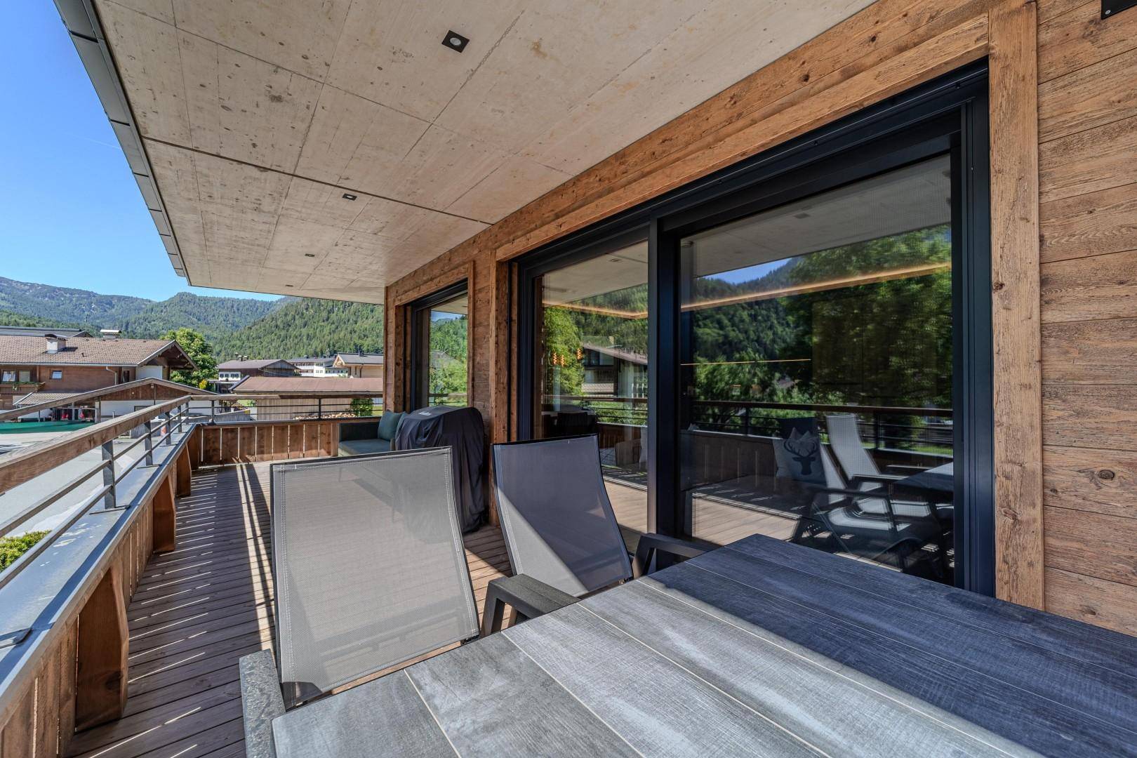 Sonniger Holzbalkon mit Esstisch und Stühlen, umgeben von grüner Berglandschaft.
