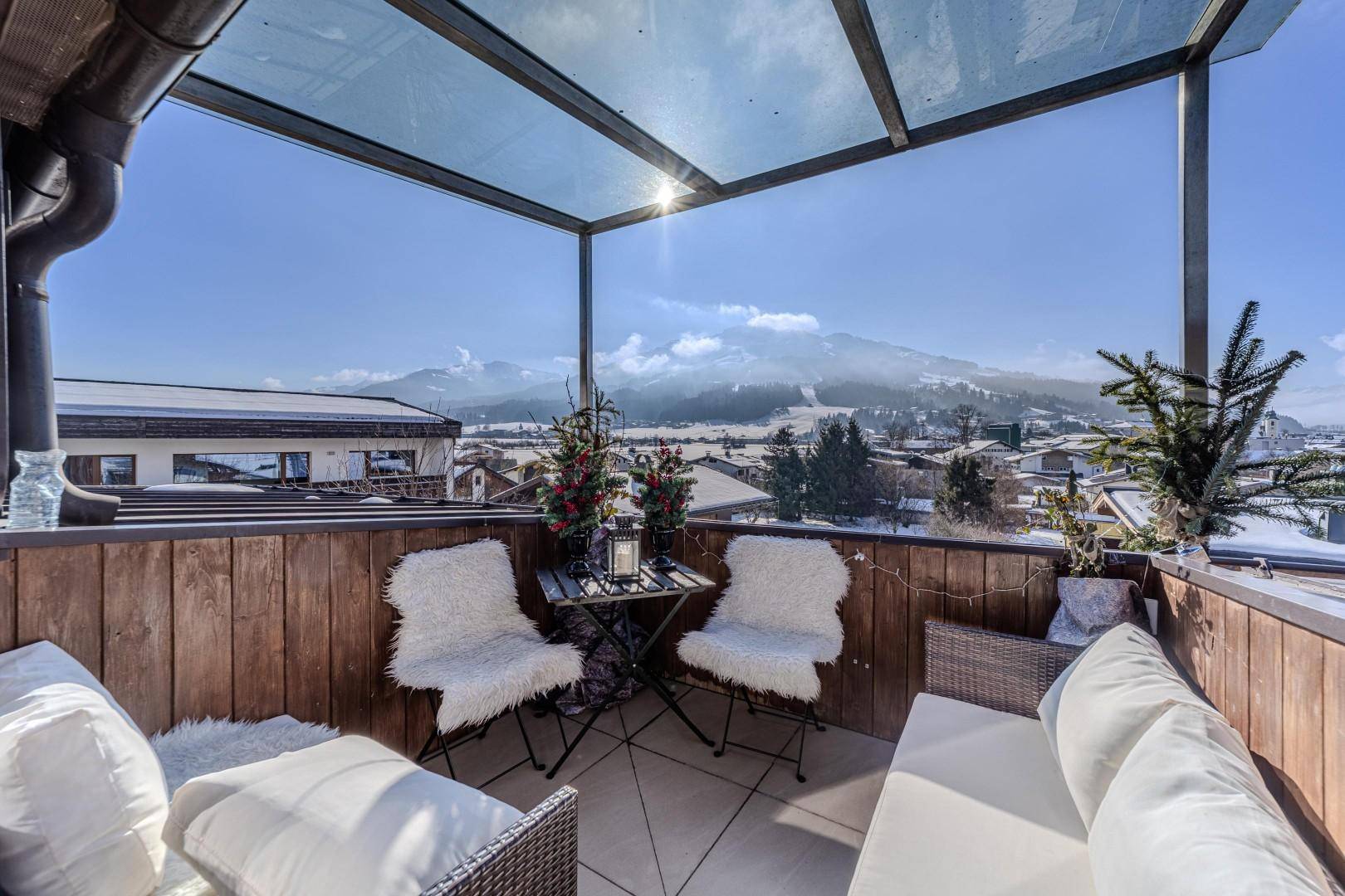 Sonniger Balkon mit Glasdach, Sitzgelegenheiten und Panoramablick auf die verschneiten Berge.