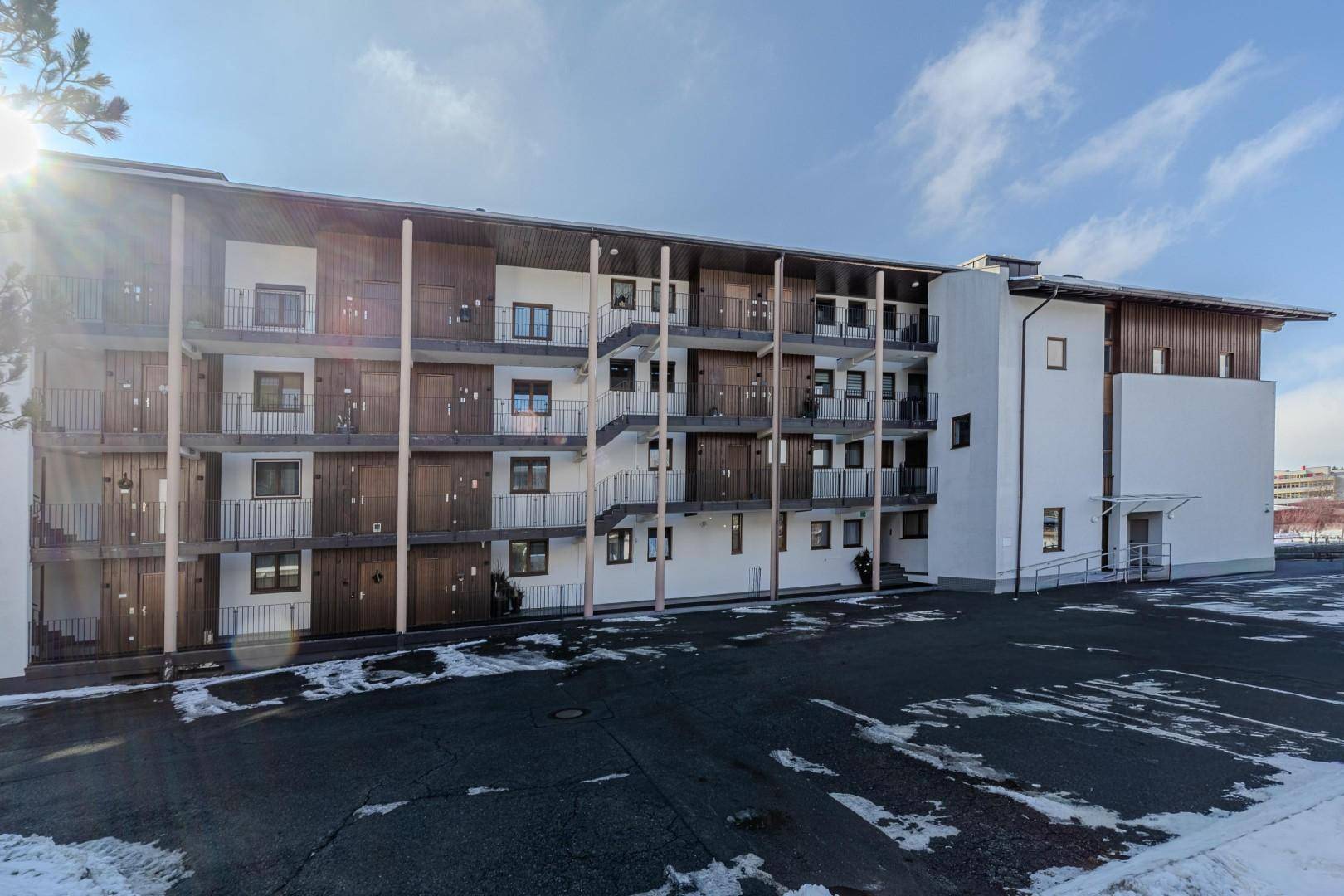 Gesamtansicht eines modernen Gebäudes mit mehreren Balkonen und einem großen Parkplatz im Vordergrund.